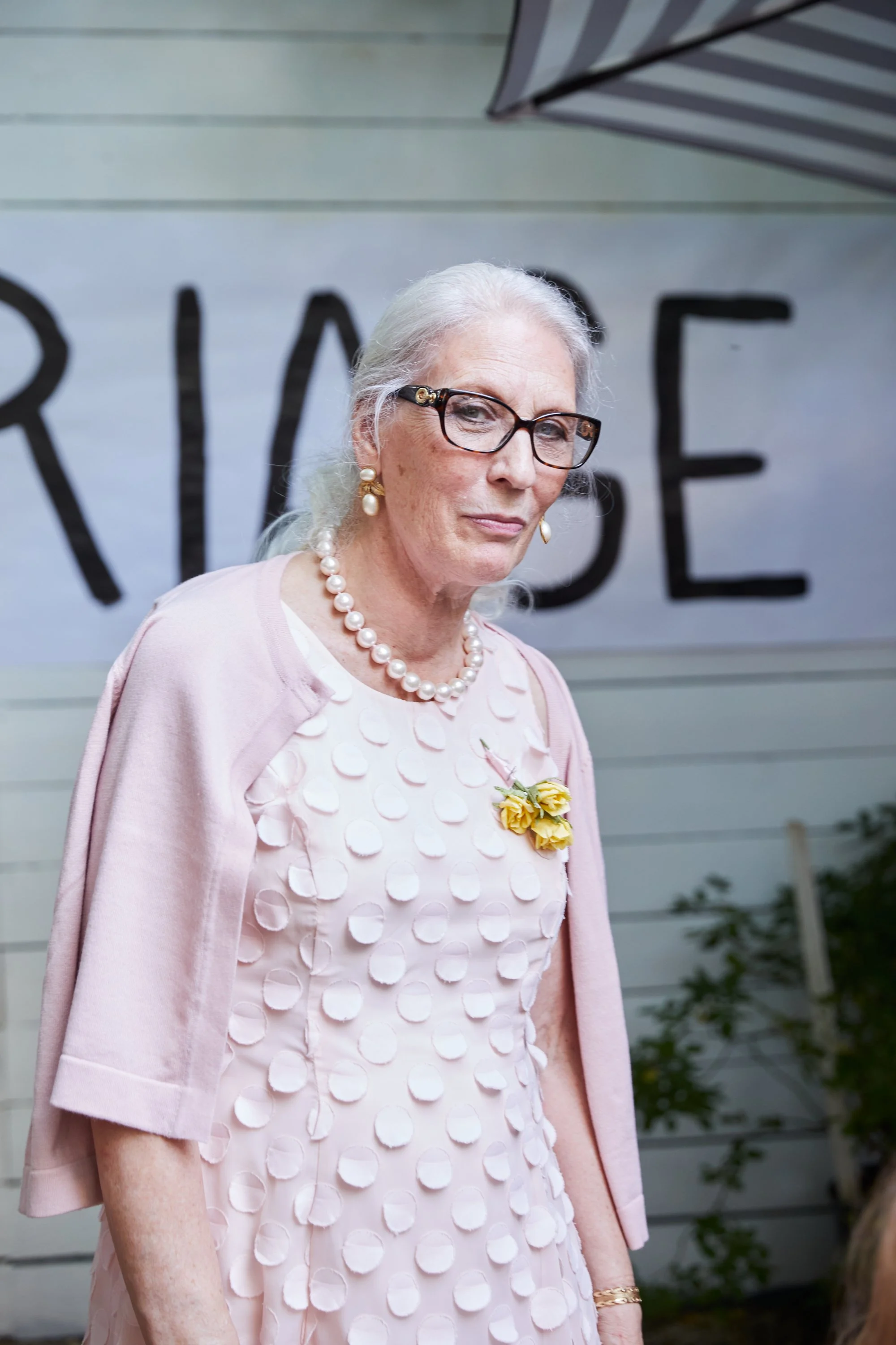 An elderly woman with gray hair, glasses, and pearl earrings wearing a light pink dress with textured circular patterns and a pink shawl, standing outdoors in front of a sign with tattered letters.