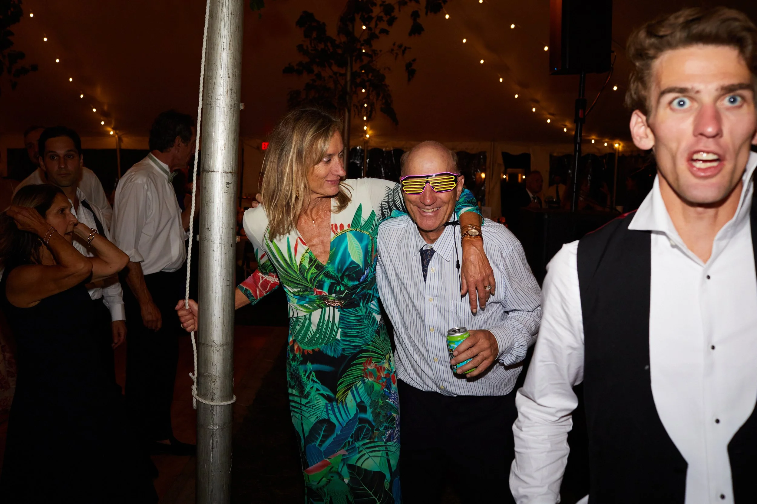 People dancing and celebrating at a party inside a decorated tent, with string lights overhead. A woman in a colorful tropical dress and a man with glasses and a drink are smiling and enjoying themselves.
