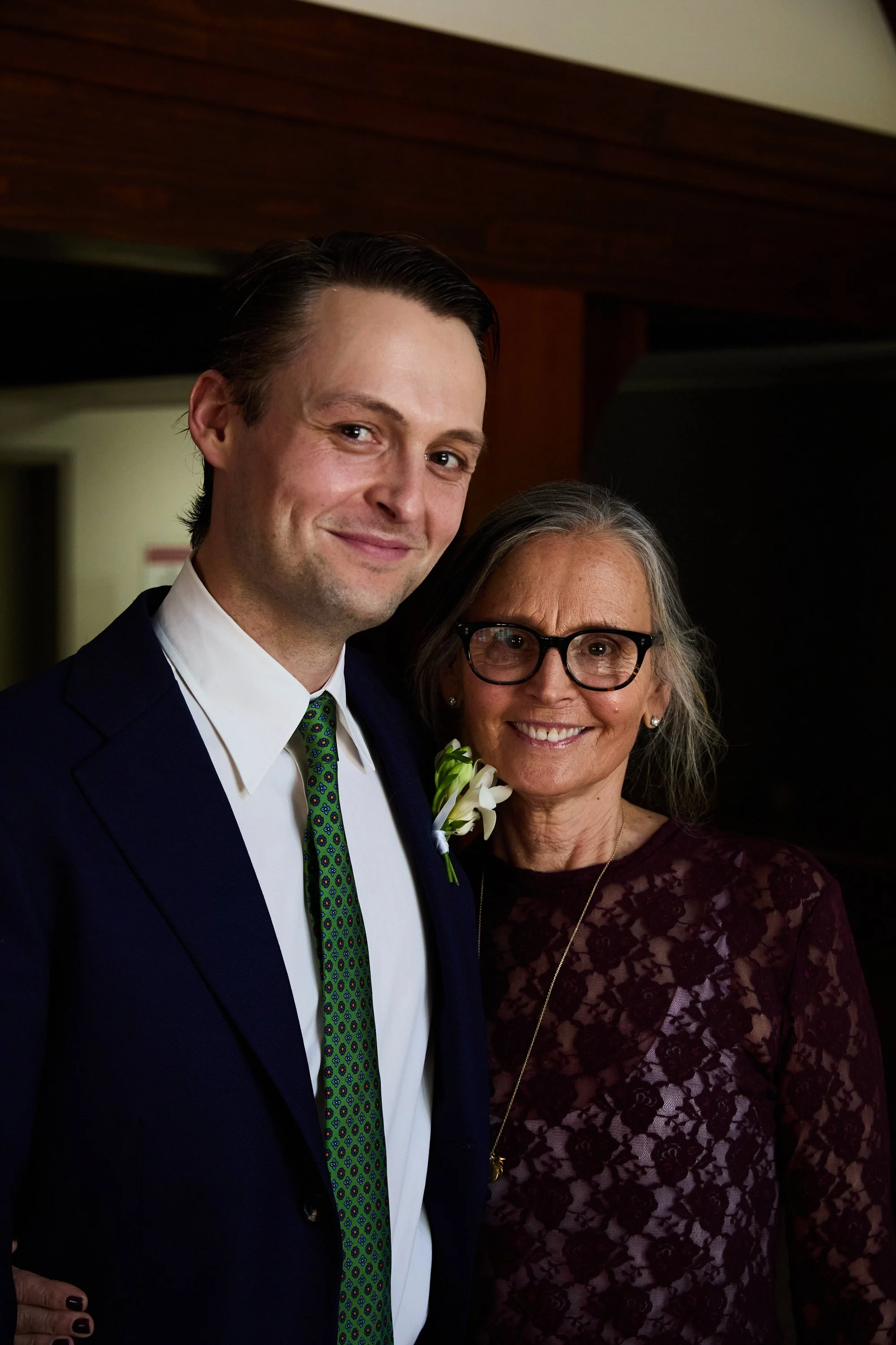 A young man in a suit with a green patterned tie standing next to an older woman with gray hair, glasses, and a burgundy lace dress, both smiling for the camera in an indoor setting.