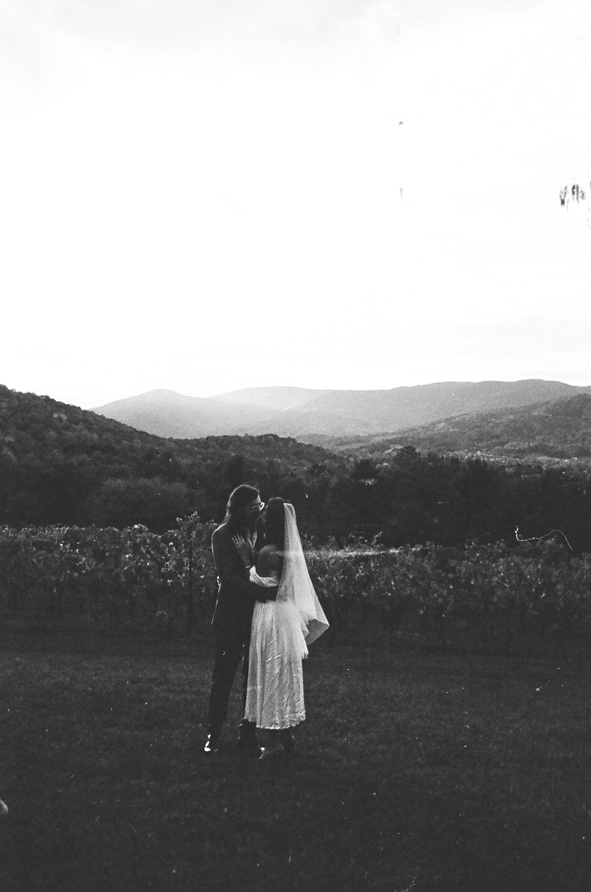 A black and white photograph of a couple in wedding attire sharing an intimate moment outdoors with a mountain range in the background at sunset or sunrise.