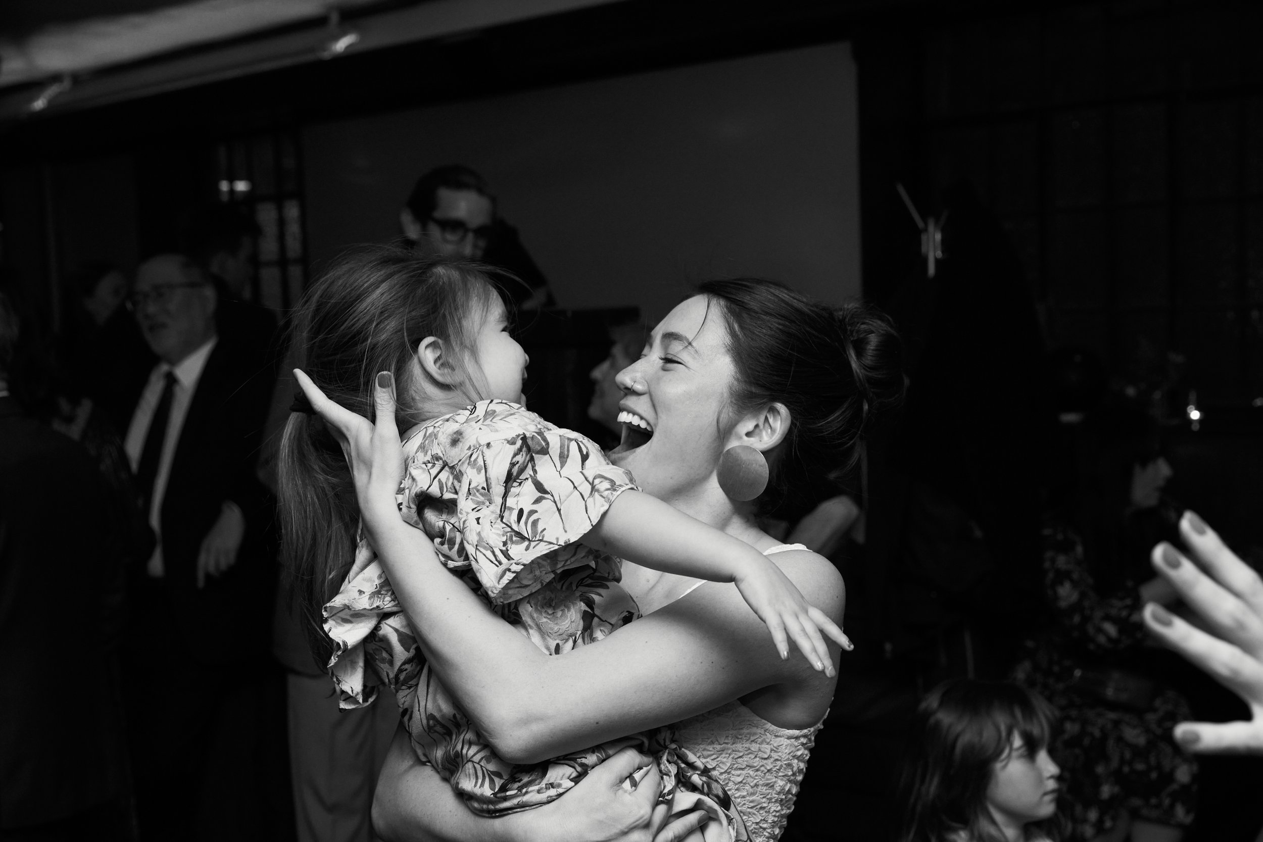 A woman with dark hair in an updo and large earrings joyfully holding a young girl with long hair in a patterned dress, at a social gathering or party.