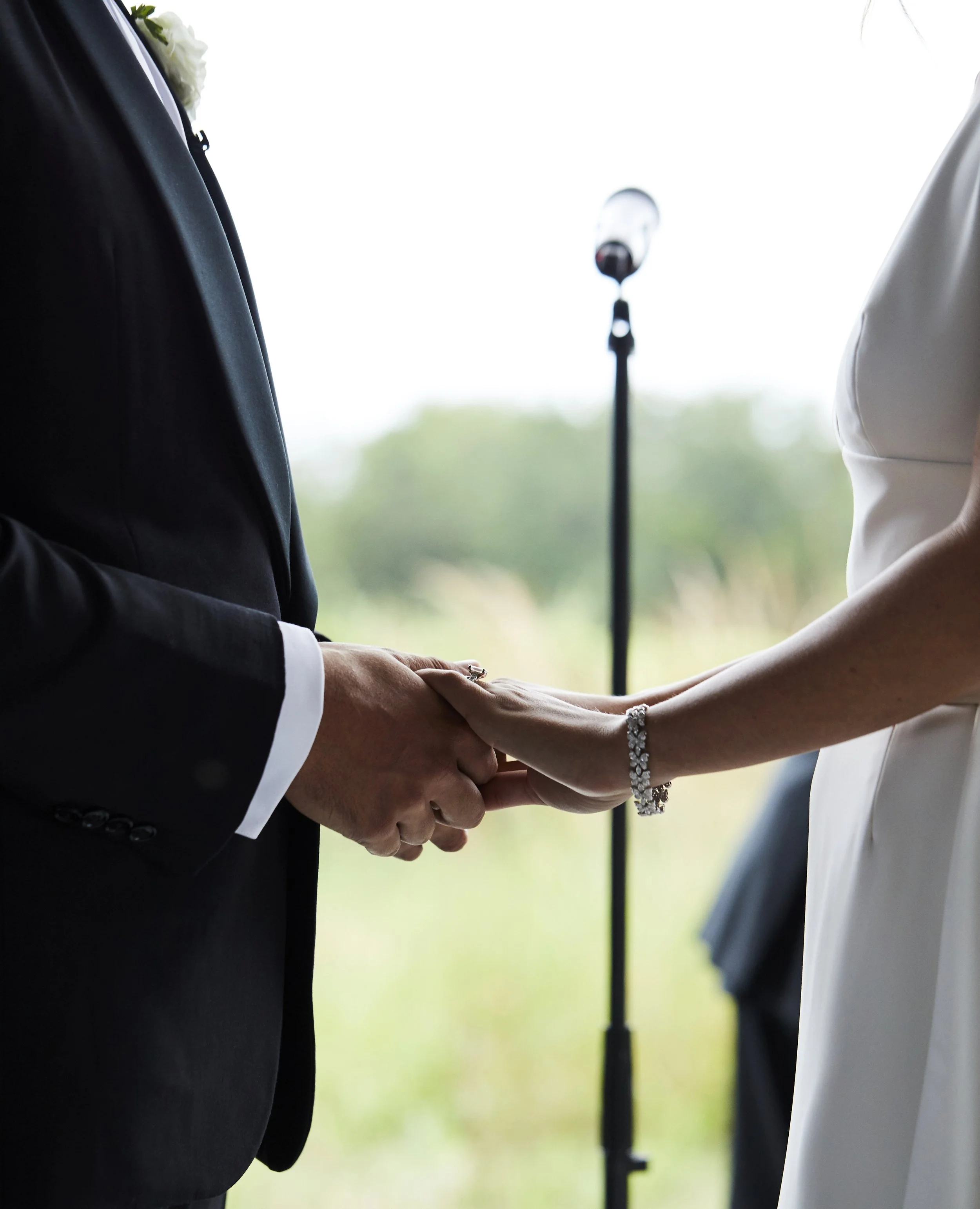 A couple holding hands during a wedding ceremony, with the groom in a black suit and the bride in a white dress, standing in front of a microphone against a blurred outdoor background.