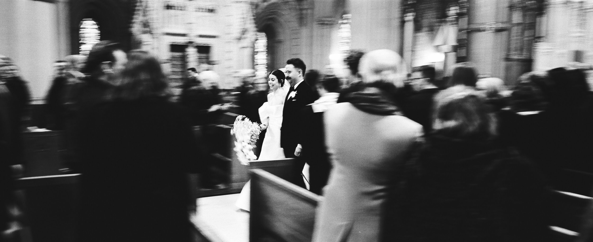 A black and white photo of a couple in wedding attire walking down the aisle in a church, surrounded by blurred guests.