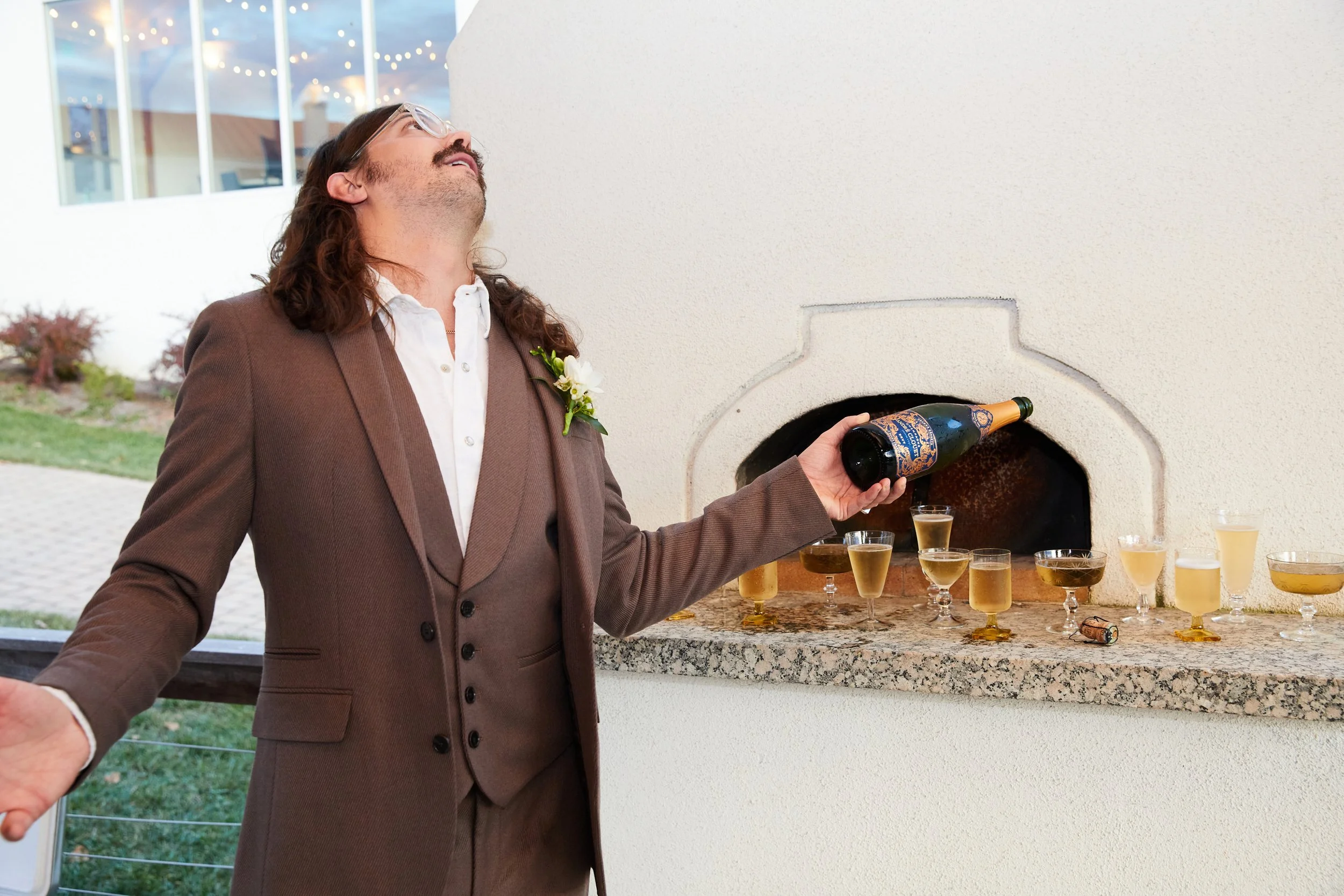 Man in a brown suit with a white shirt and boutonniere pouring champagne from a bottle into glasses at a celebration or event.