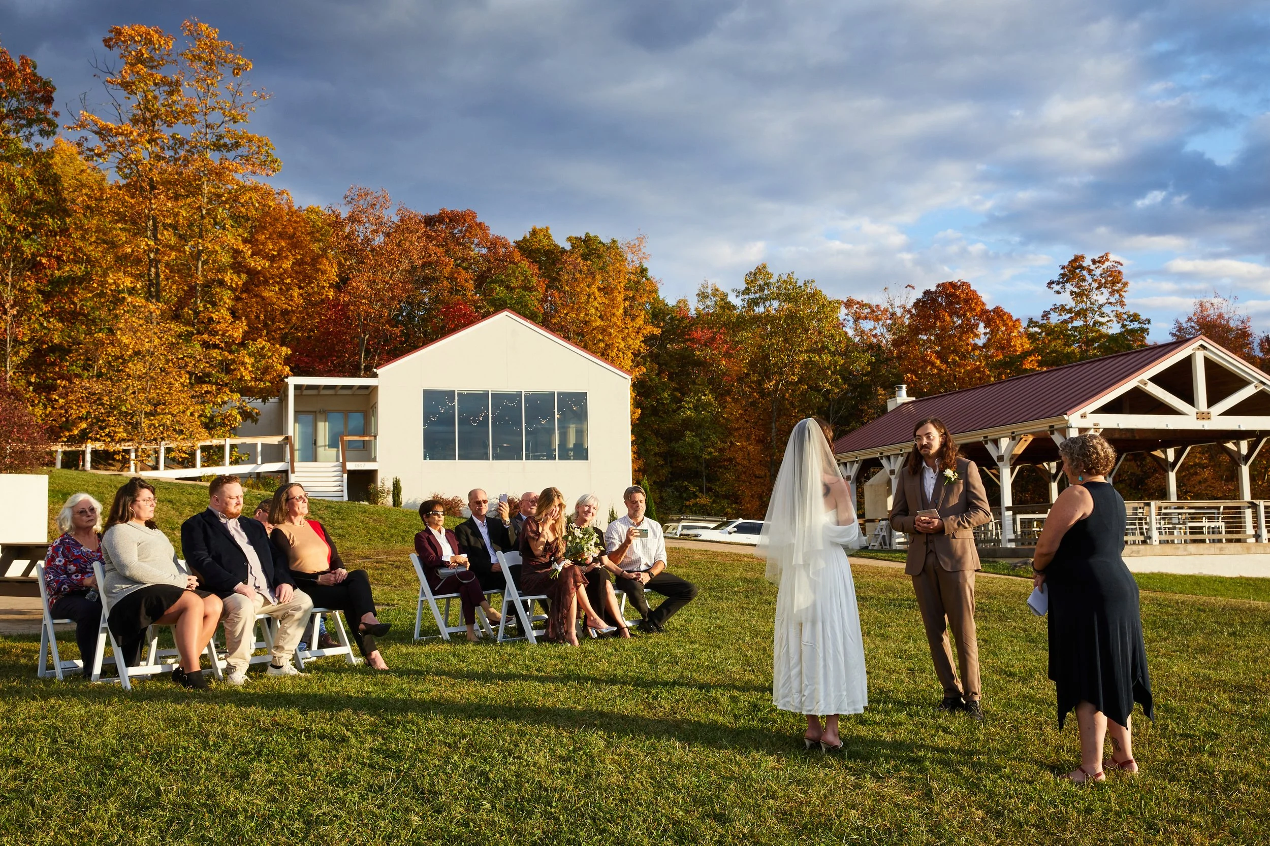 Couple exchanging vows during outdoor wedding ceremony on grassy lawn, guests seated on white chairs, autumn trees and modern building in background.
