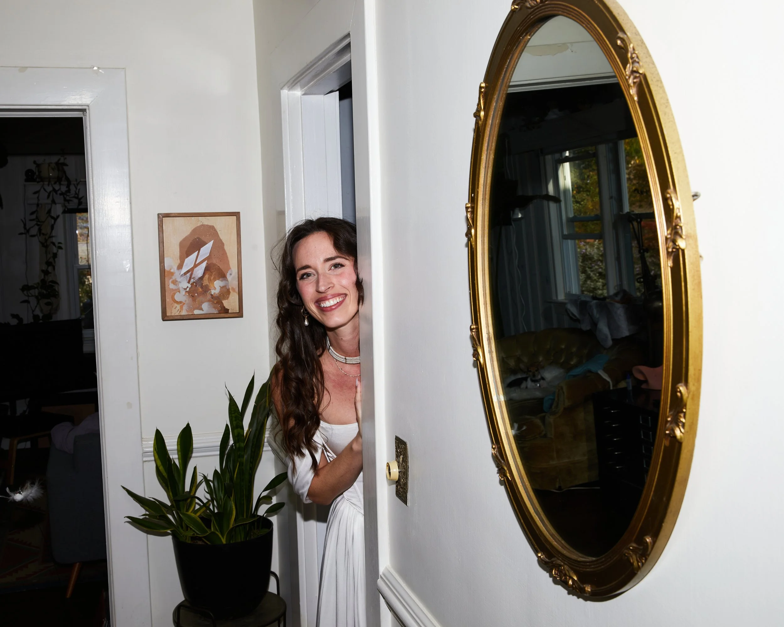 A woman with long wavy brown hair and wearing a white dress and jewelry peeking out from behind a wall and smiling at the camera inside a home. A gold-framed mirror and a potted plant are visible in the background.