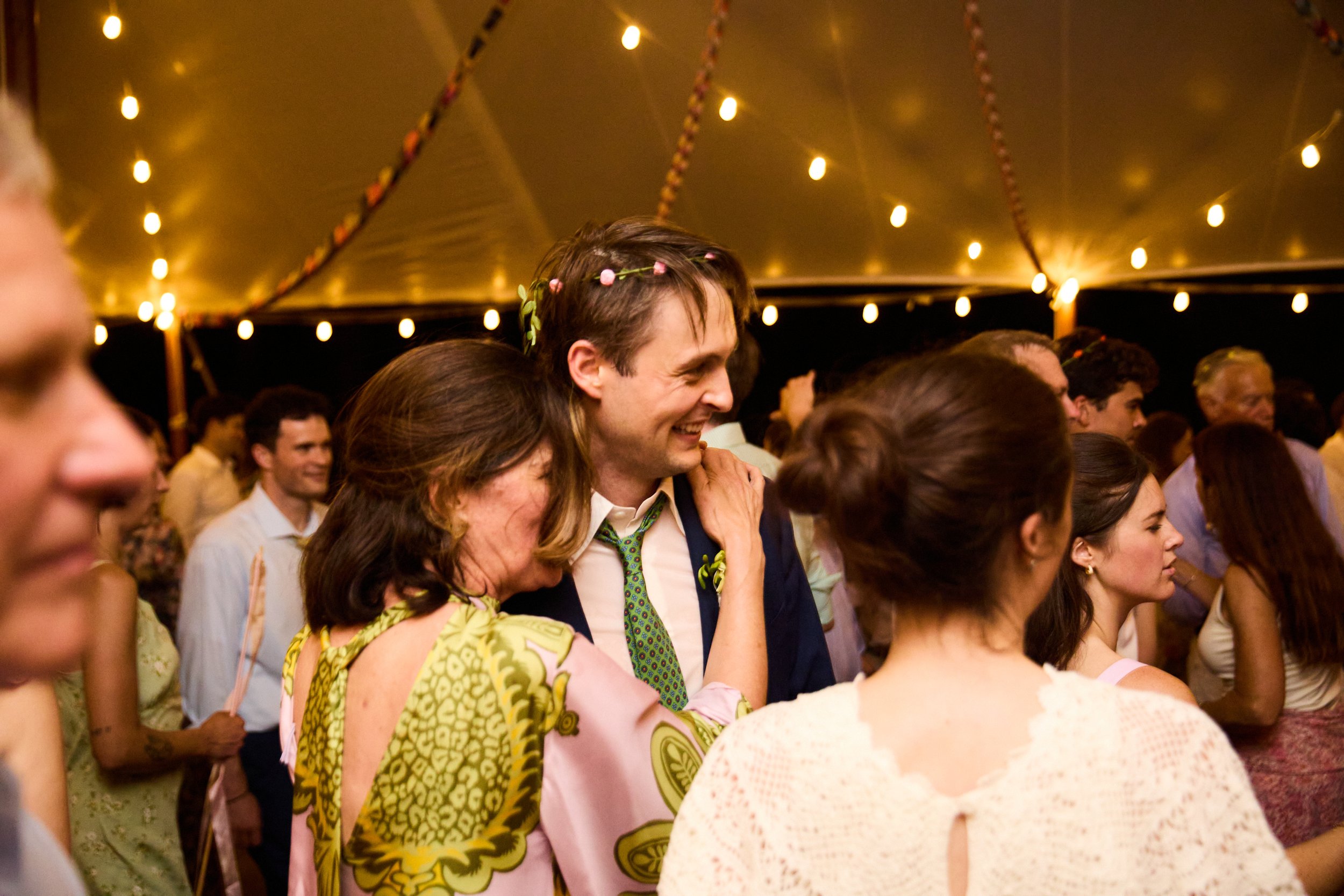People dancing and smiling at a celebration or party under a decorated tent with string lights.
