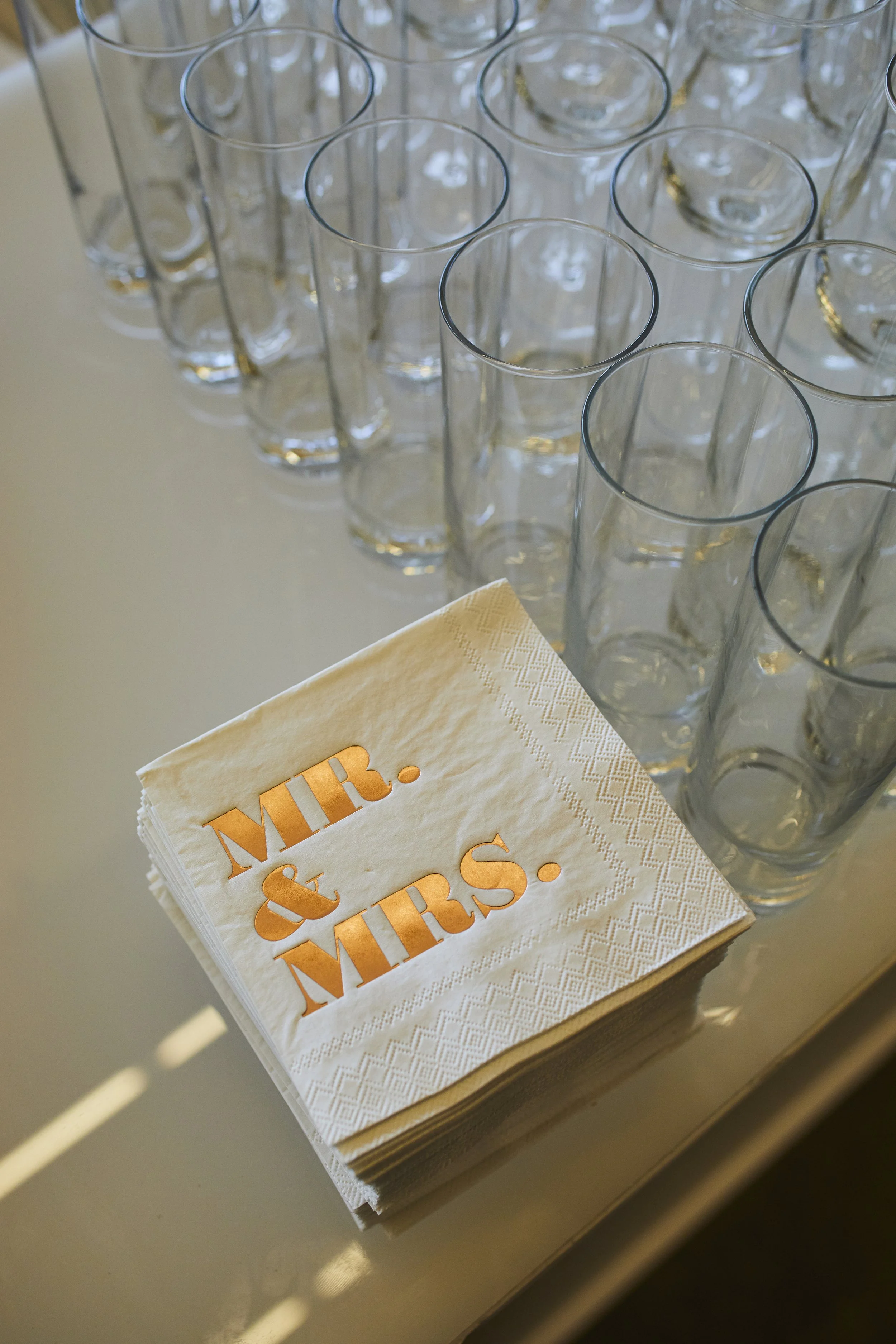A stack of napkins with 'Mr. & Mrs.' printed in metallic gold, placed next to a row of empty tall drinking glasses on a white table.