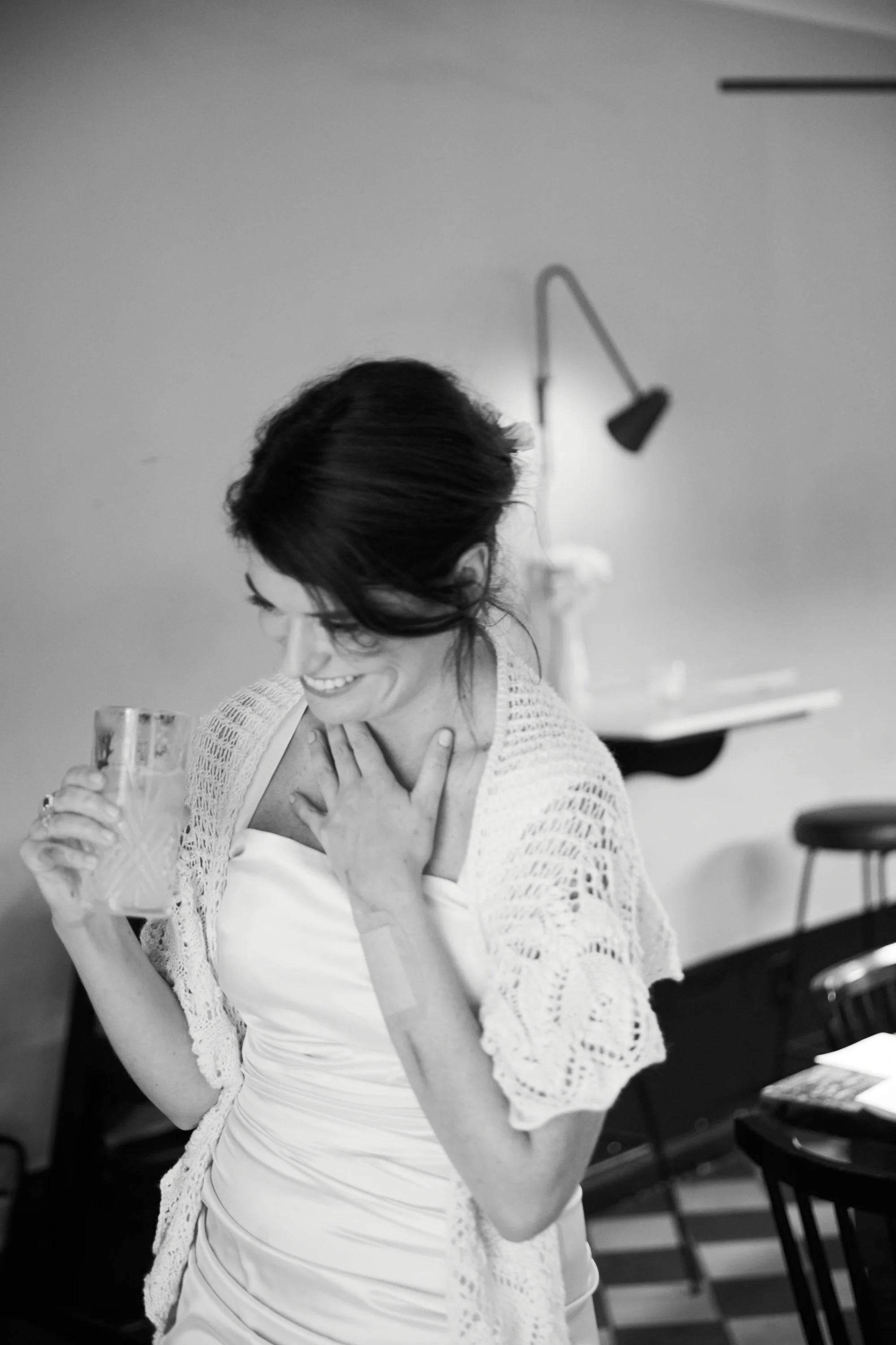 A woman in a white dress and lace shawl holding a glass, smiling with her hand on her chest, in a room with chairs and a wall lamp.