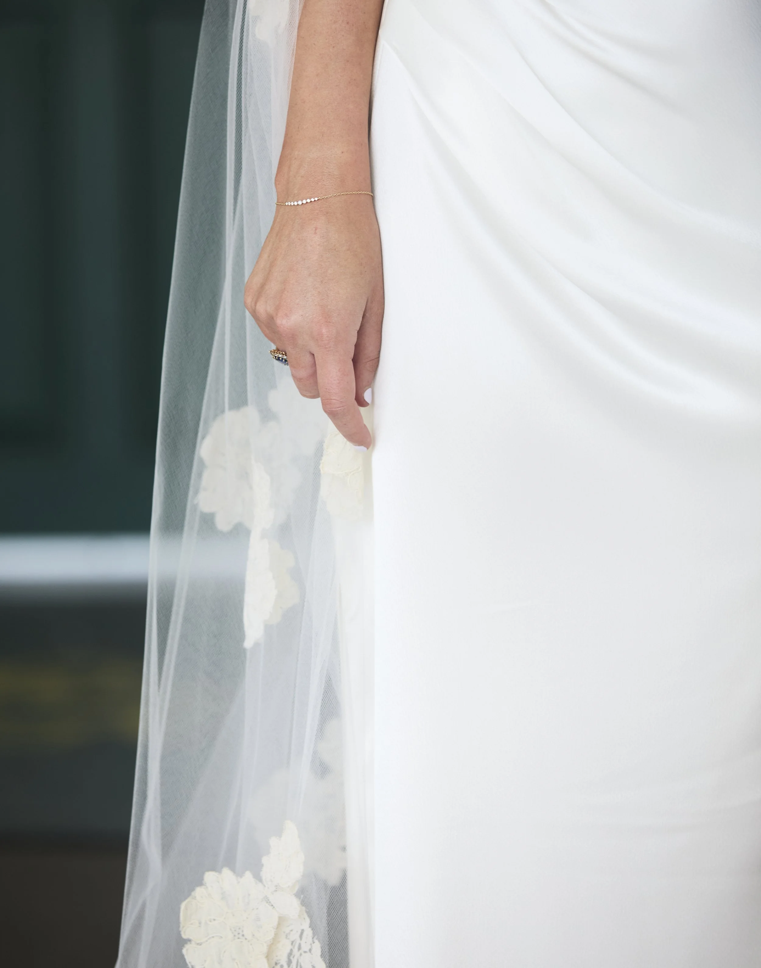 Close-up of a bride's hand resting on her white wedding dress, with a delicate bracelet and a ring on her finger, and sheer lace fabric with floral embroidery.