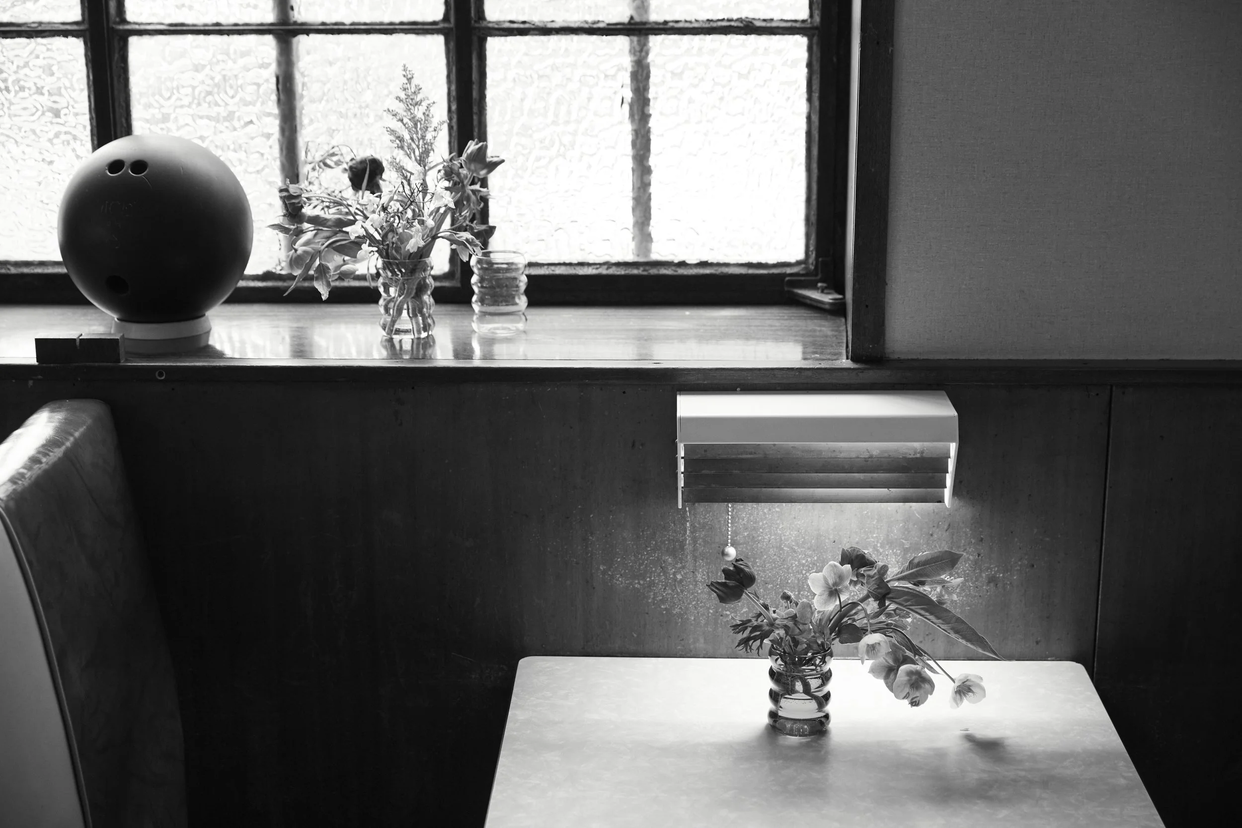 Black and white photo of an interior scene with a window sill holding a spherical speaker, a small bouquet of flowers in a glass vase, and a jar, with another vase of flowers on a table below near a vent, all with soft light coming through the window