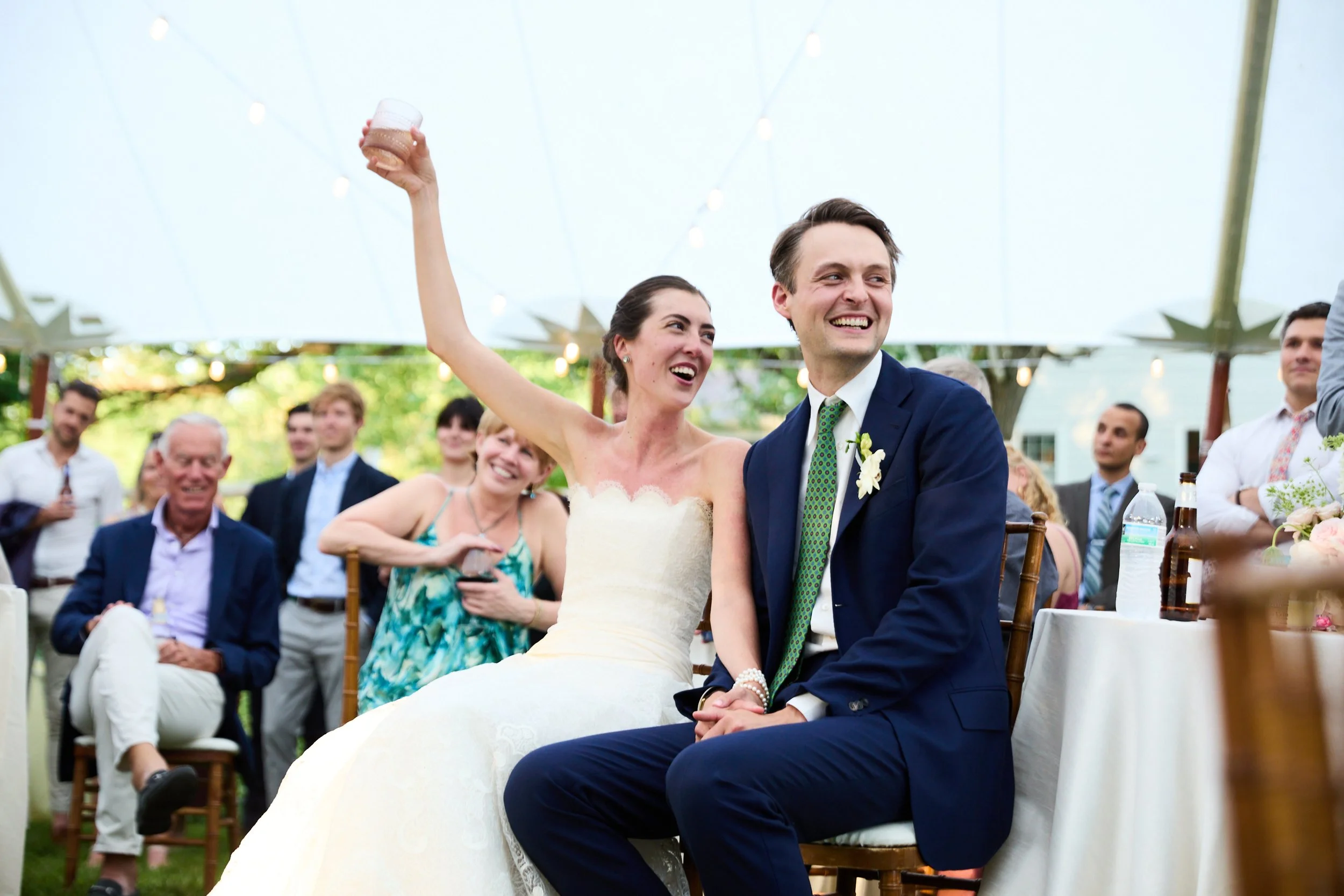 Bride and groom smiling at their wedding reception, with guests in the background, outdoors with string lights overhead.