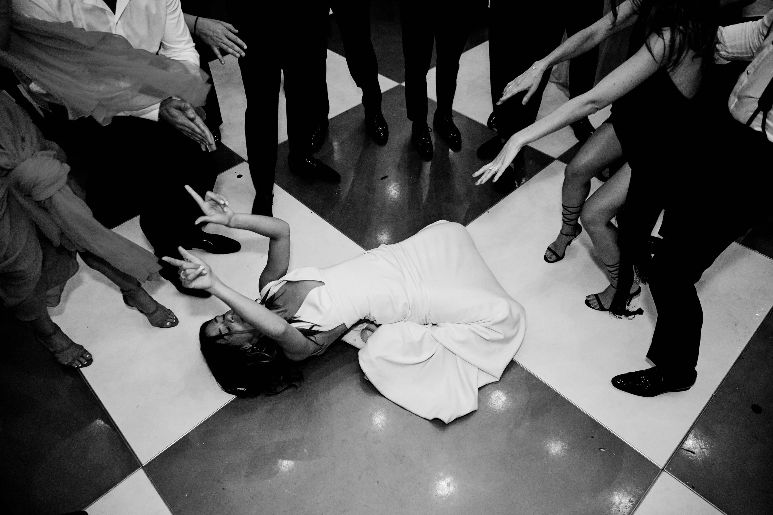 A woman in a white dress is lying on the floor, surrounded by a group of people at an event. She appears to be posing or dancing, with her arms raised and her eyes closed. The floor is checkered with black and white tiles.