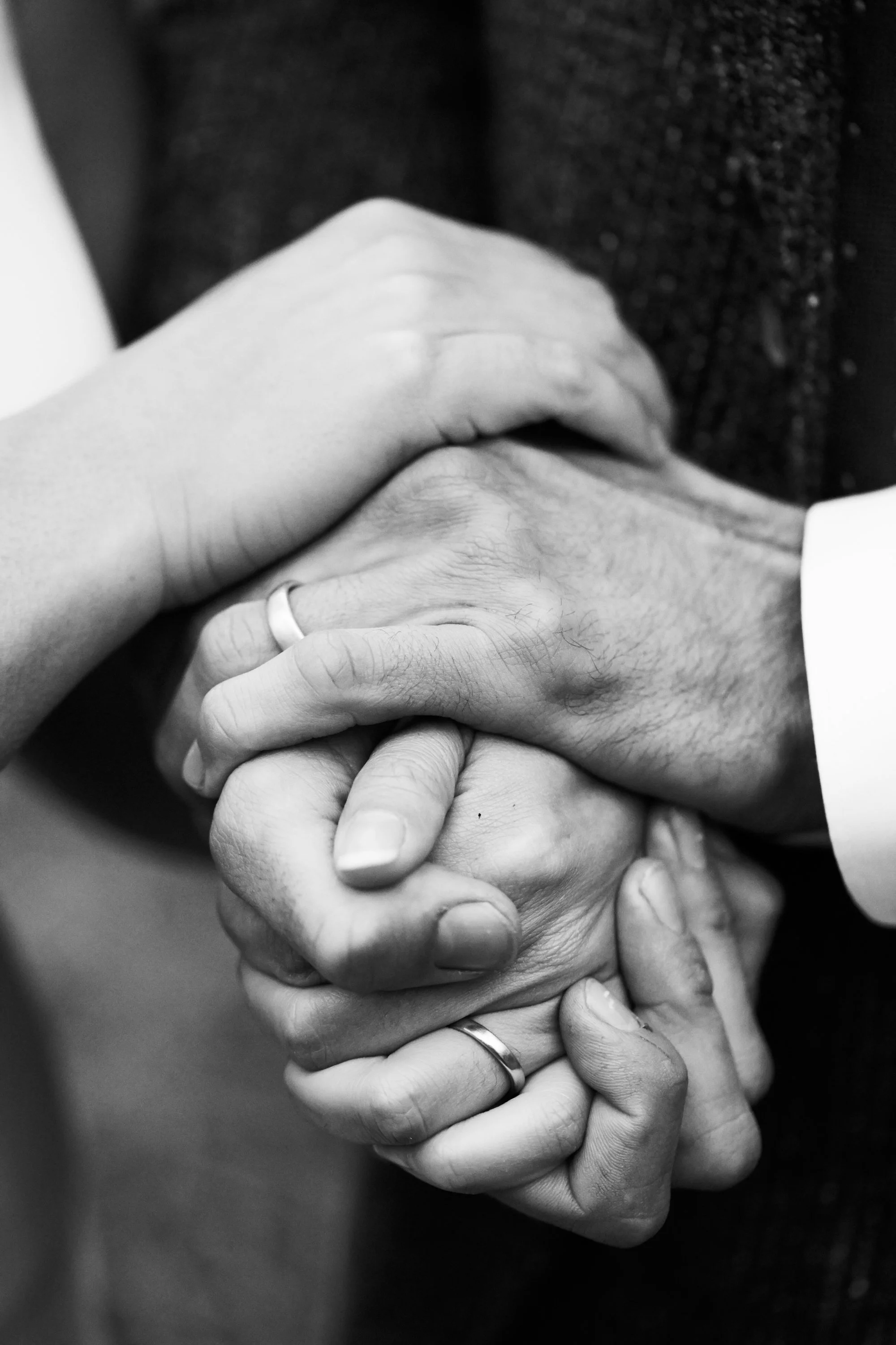 Close-up of four hands holding each other, two with wedding rings, in black and white.
