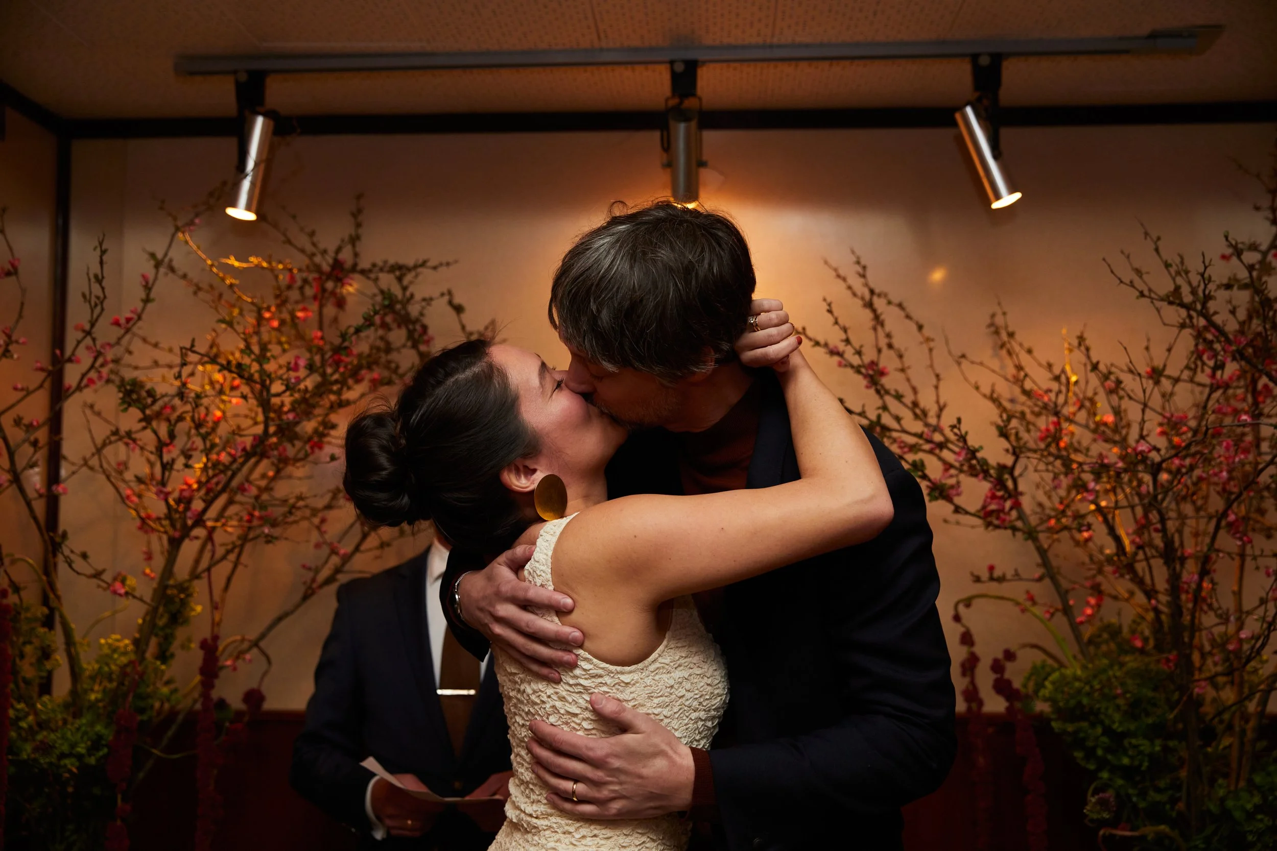 A couple kisses passionately during a wedding ceremony, with a woman wearing a cream lace dress and large gold earrings, and a man in a dark suit, in front of a backdrop of flowering branches, with an officiant holding a book in the background.