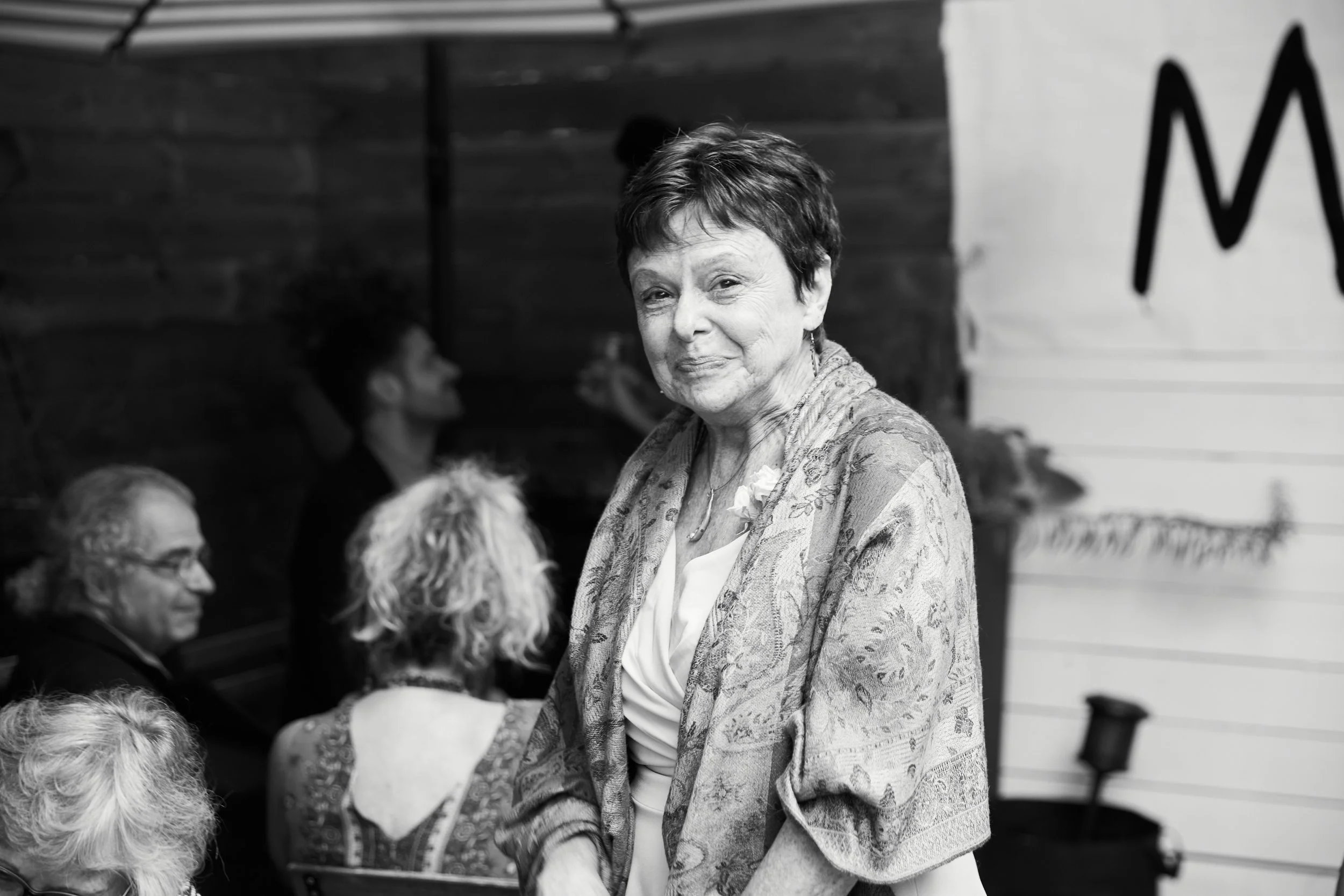 An older woman with short hair and a patterned shawl stands in front of a group of seated people, smiling slightly. The background has a wooden wall and a banner with part of a letter "M."