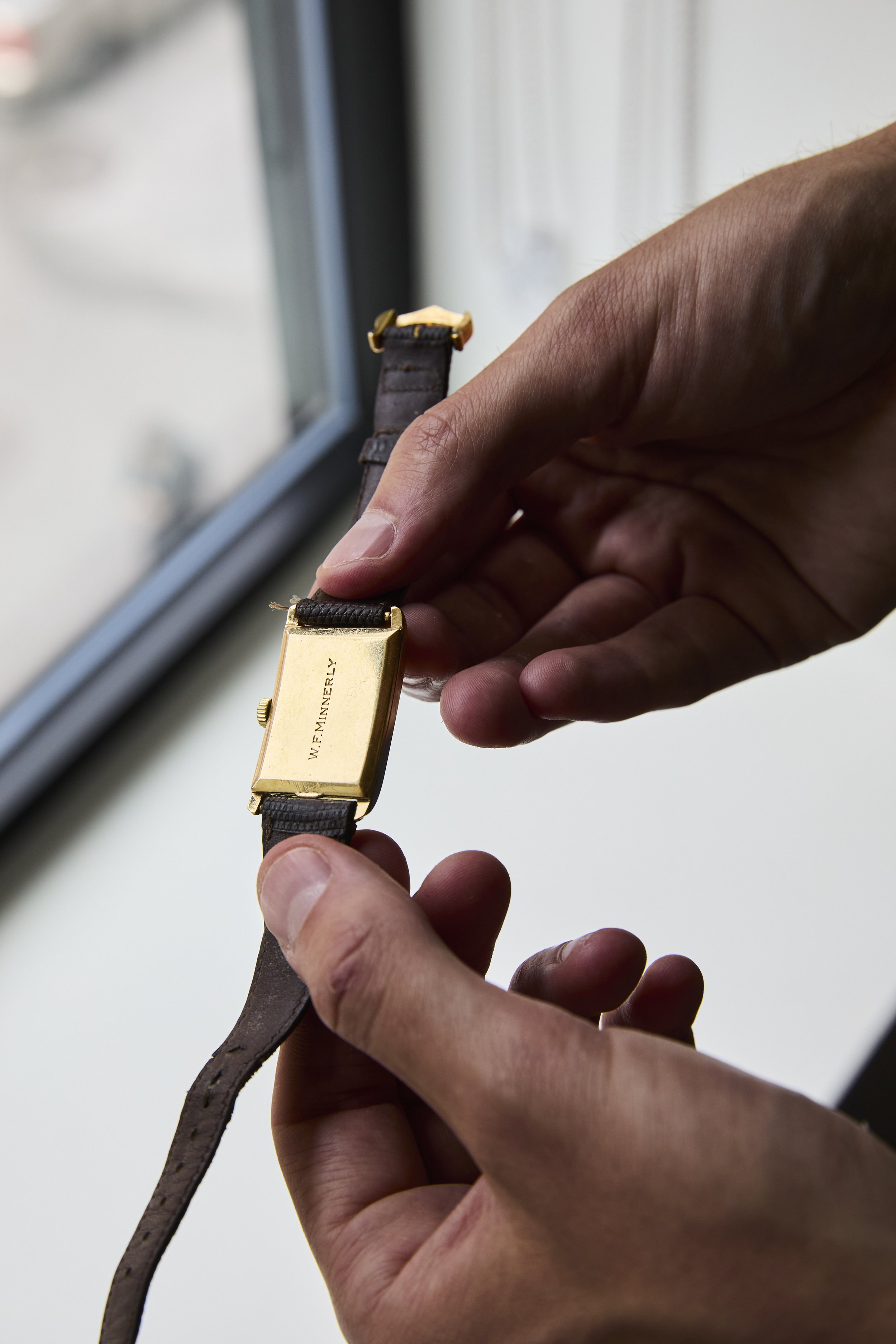A person holding a gold vintage wristwatch with a brown leather strap near a window.