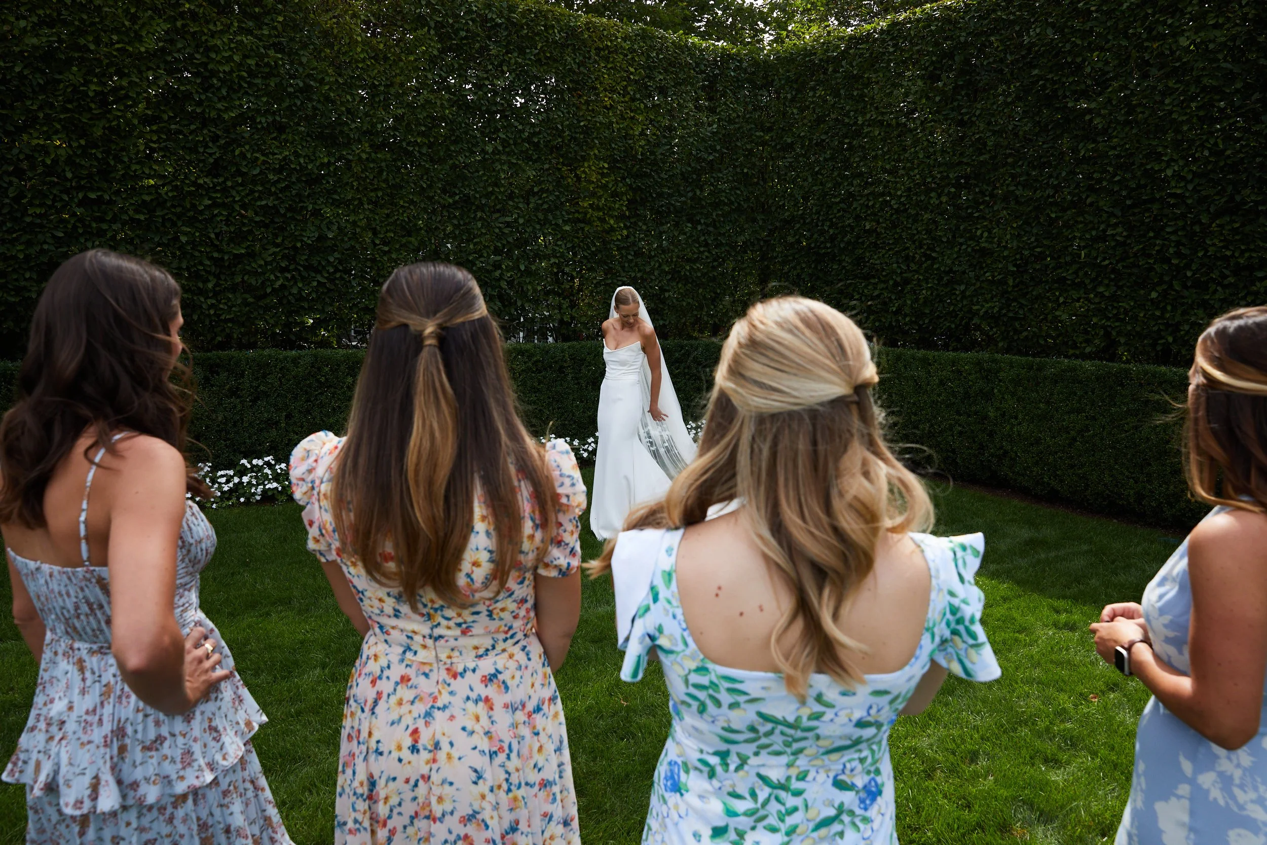 A bride in a white wedding dress holding her veil stands on a green lawn, surrounded by four women in colorful dresses, with a tall hedge in the background.