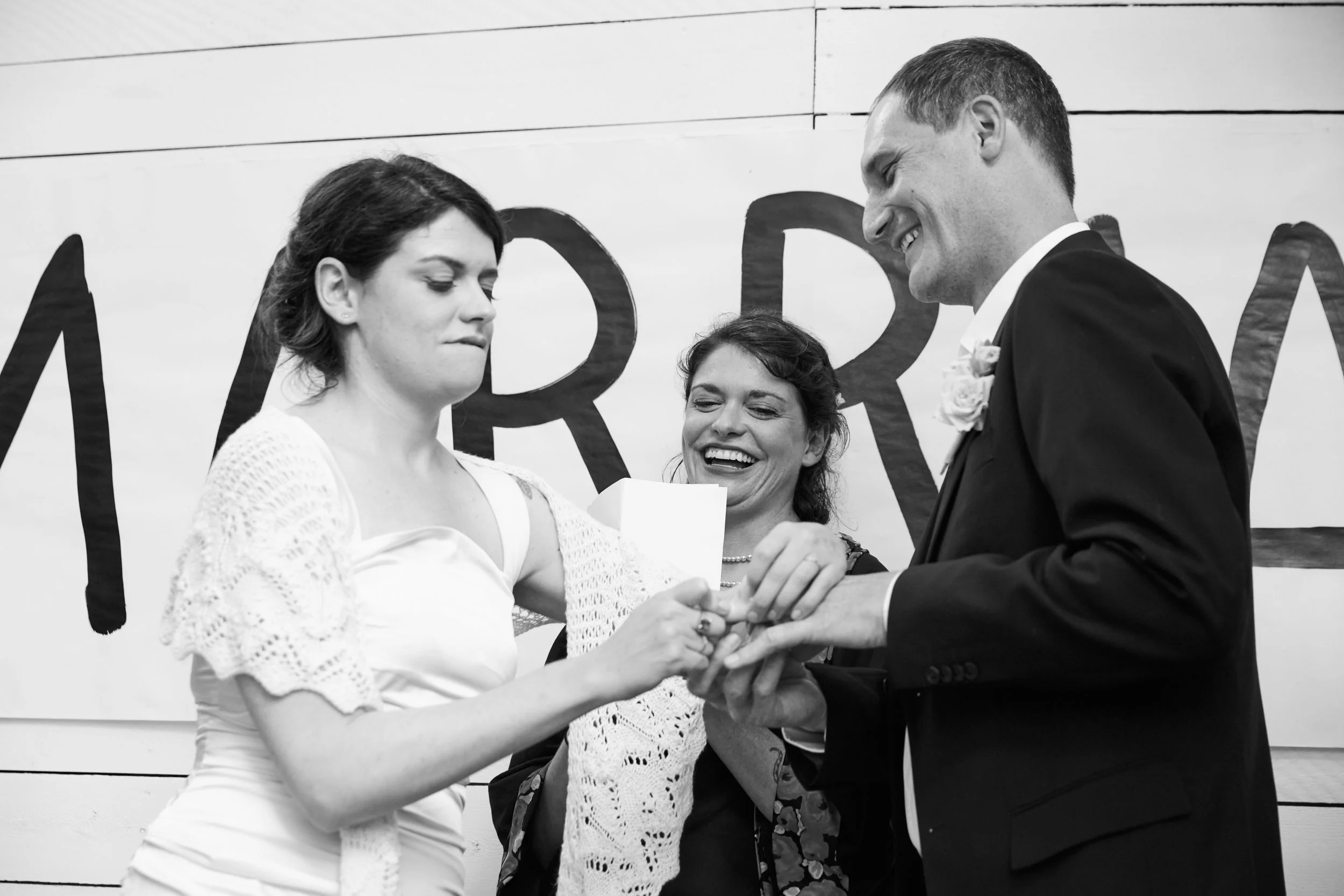 A woman in a white dress is exchanging rings with a man in a suit during a wedding ceremony, with a woman in a dark dress smiling behind them.