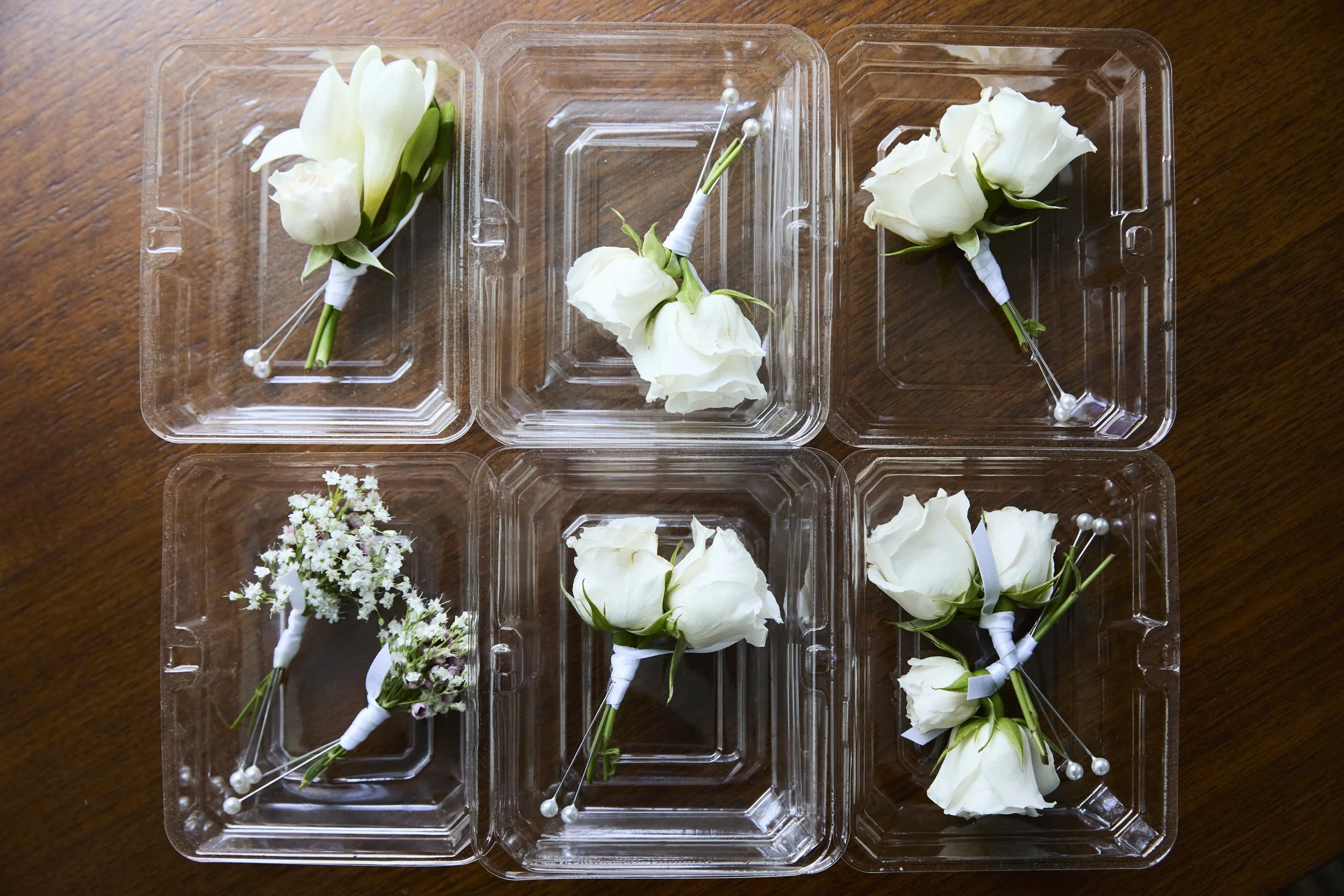 Six plastic containers with white flower boutonnieres and corsages on a wooden surface.