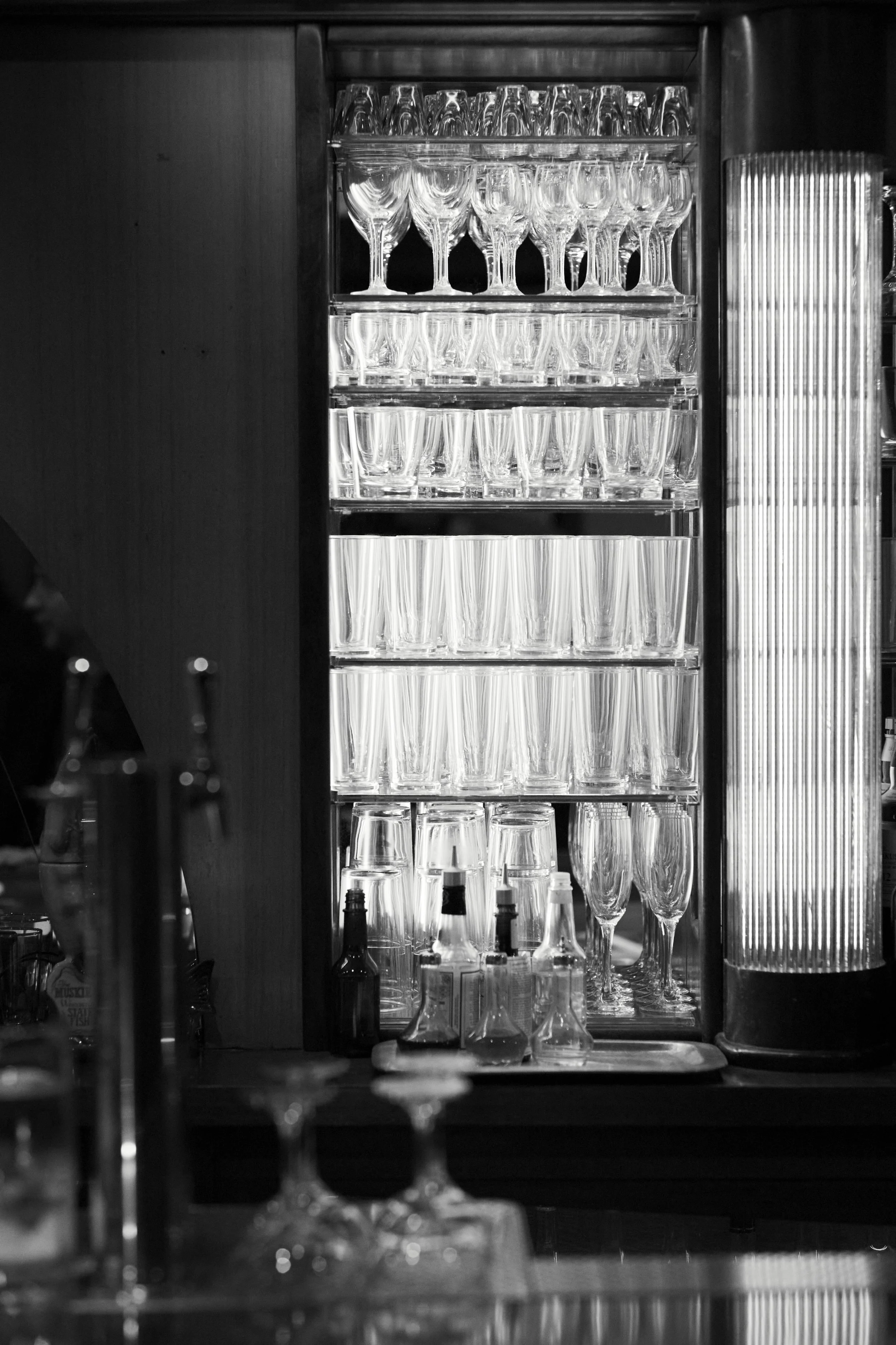 Black and white photo of a cabinet filled with glasses, including wine glasses, tumblers, and bottled beverages, in a bar setting.