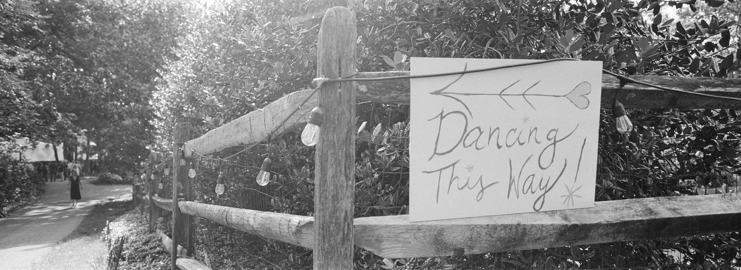 A wooden fence with a handmade sign that reads 'Dancing This Way!' hanging on it, decorated with a heart and star illustrations, along with string lights. In the background, a park pathway with trees and a person walking.