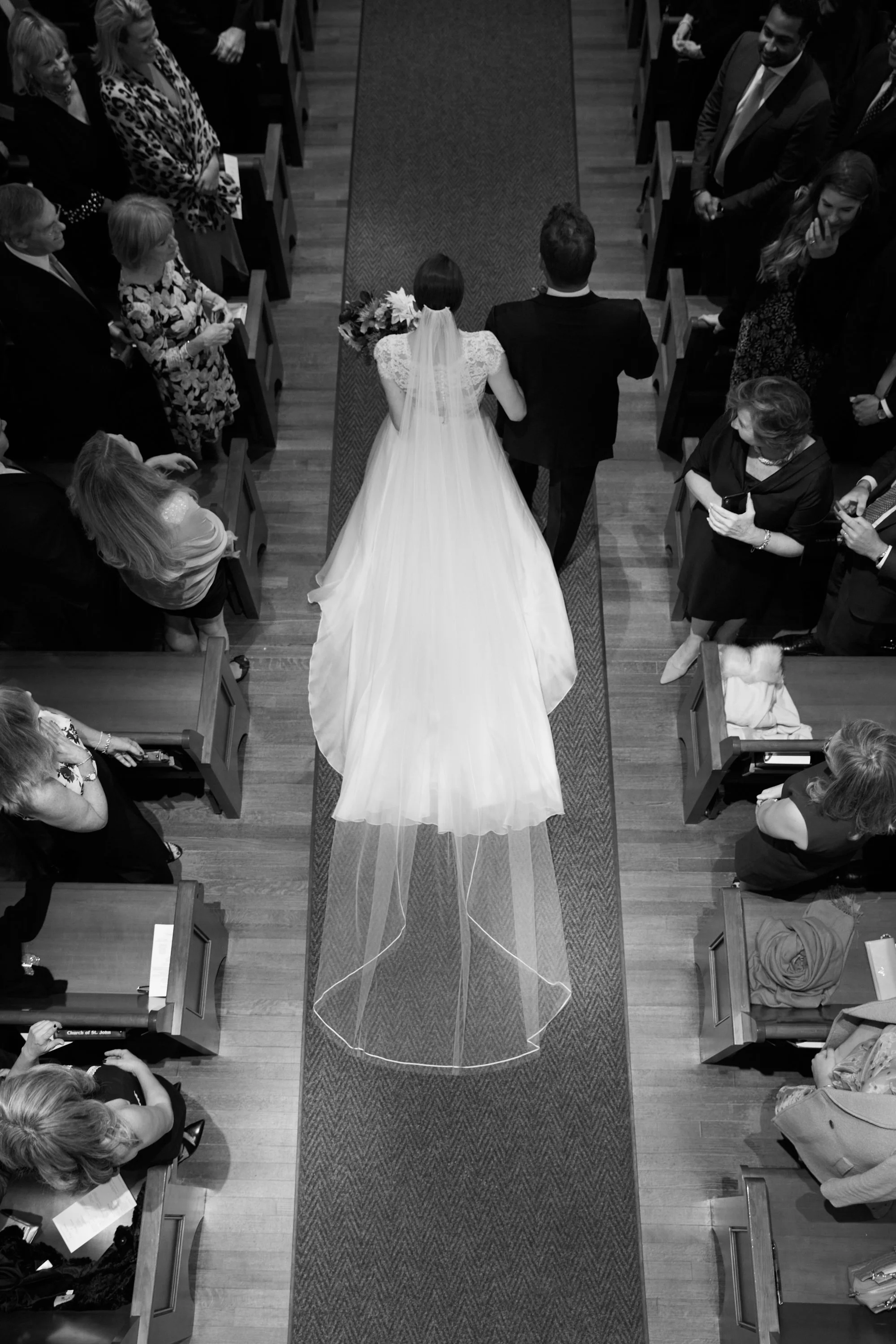 A bride in a wedding dress and a groom in a suit walking down the aisle of a church, surrounded by guests seated in pews, during a wedding ceremony.