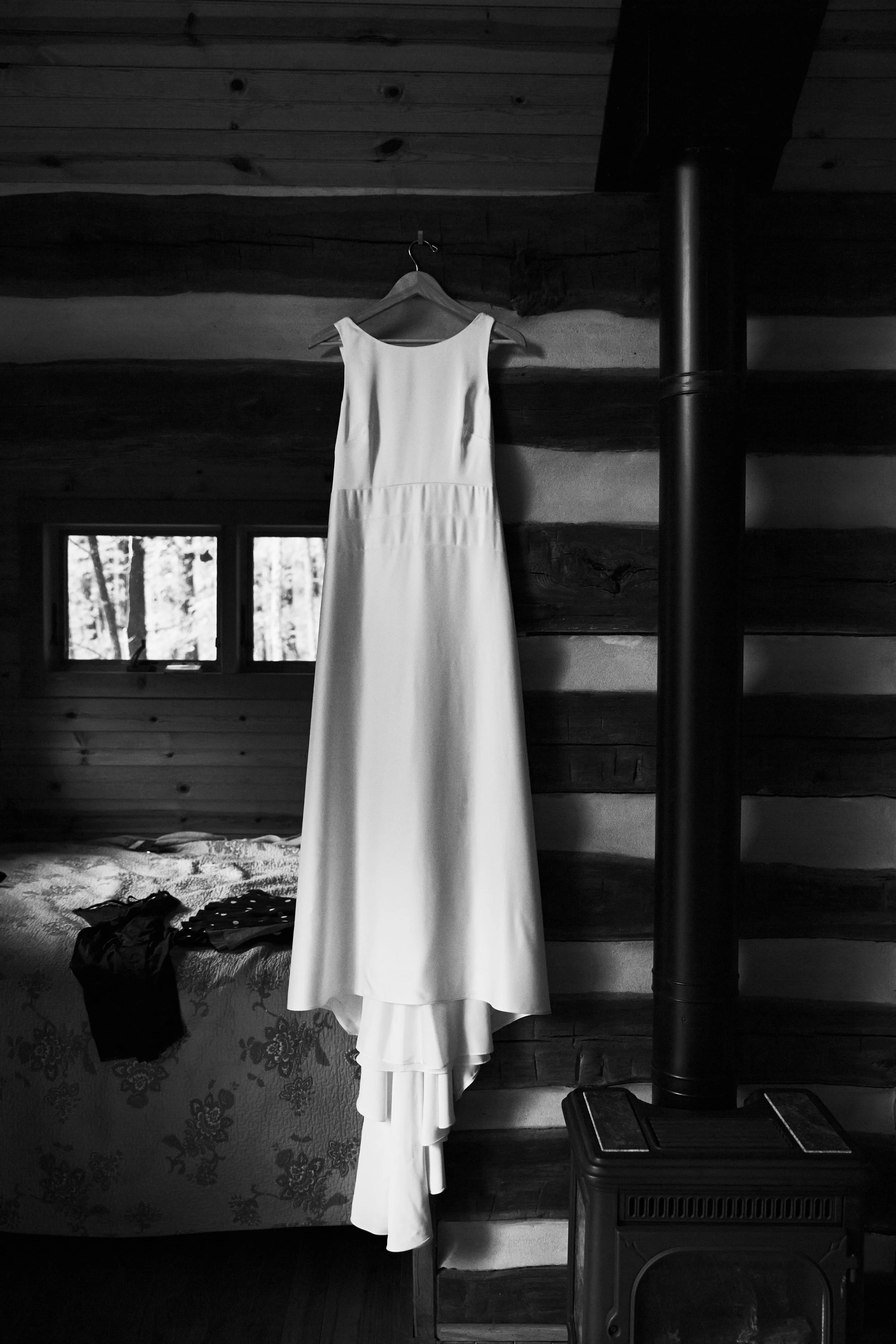 A white wedding dress hanging on a wooden wall in a rustic cabin, with a window in the background and a stove in the corner.
