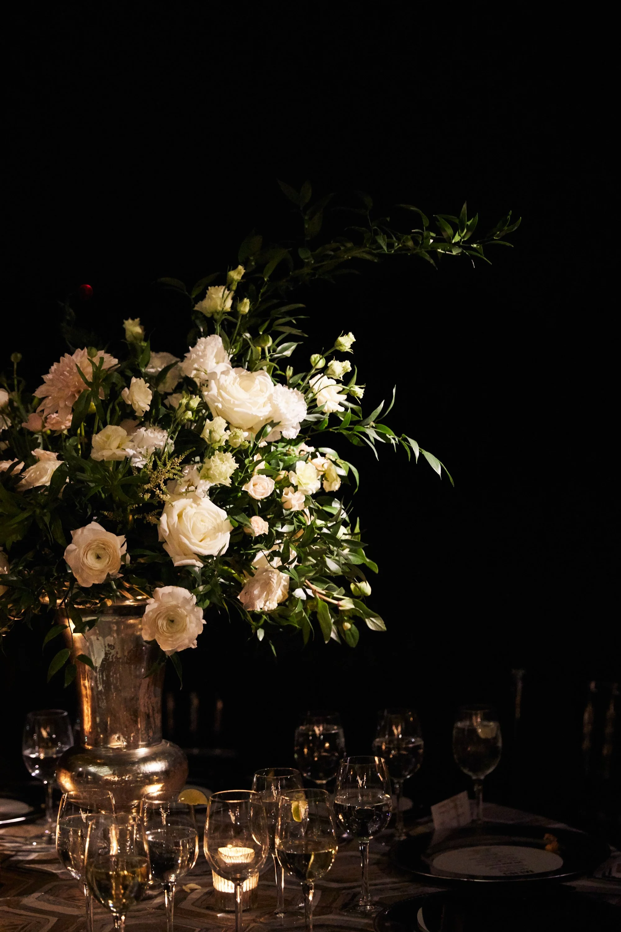A tall bouquet of white roses, peonies, and other flowers in a metallic vase, arranged on a dark table with several wine glasses, some filled with white wine, and a candle.