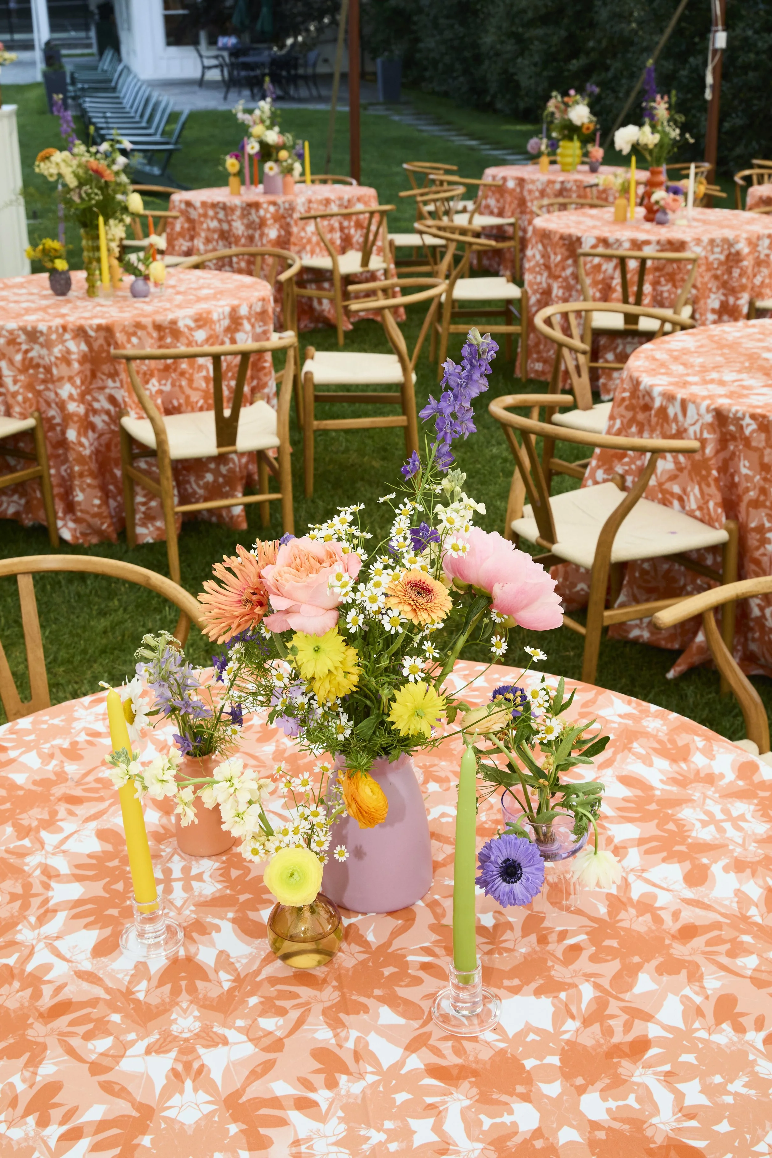 Flower centerpiece with pink, yellow, purple, and white flowers on a table with coral-colored leaf-patterned tablecloth, surrounded by chairs at an outdoor event.