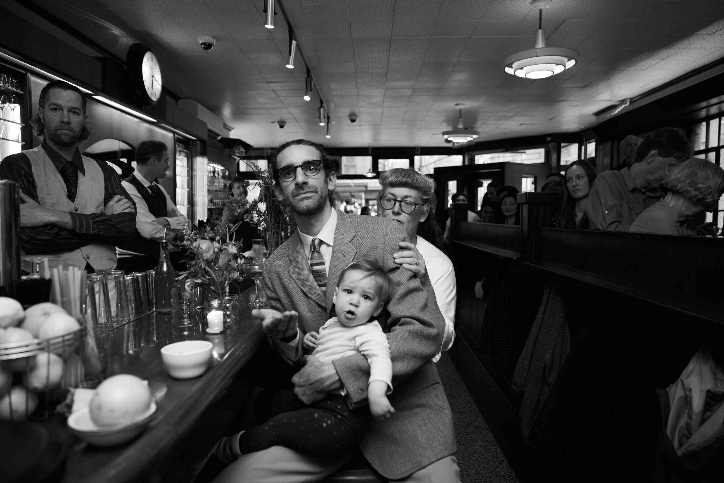 A black and white photo of a busy restaurant with a man holding a young girl in the foreground, surrounded by other diners and waitstaff.