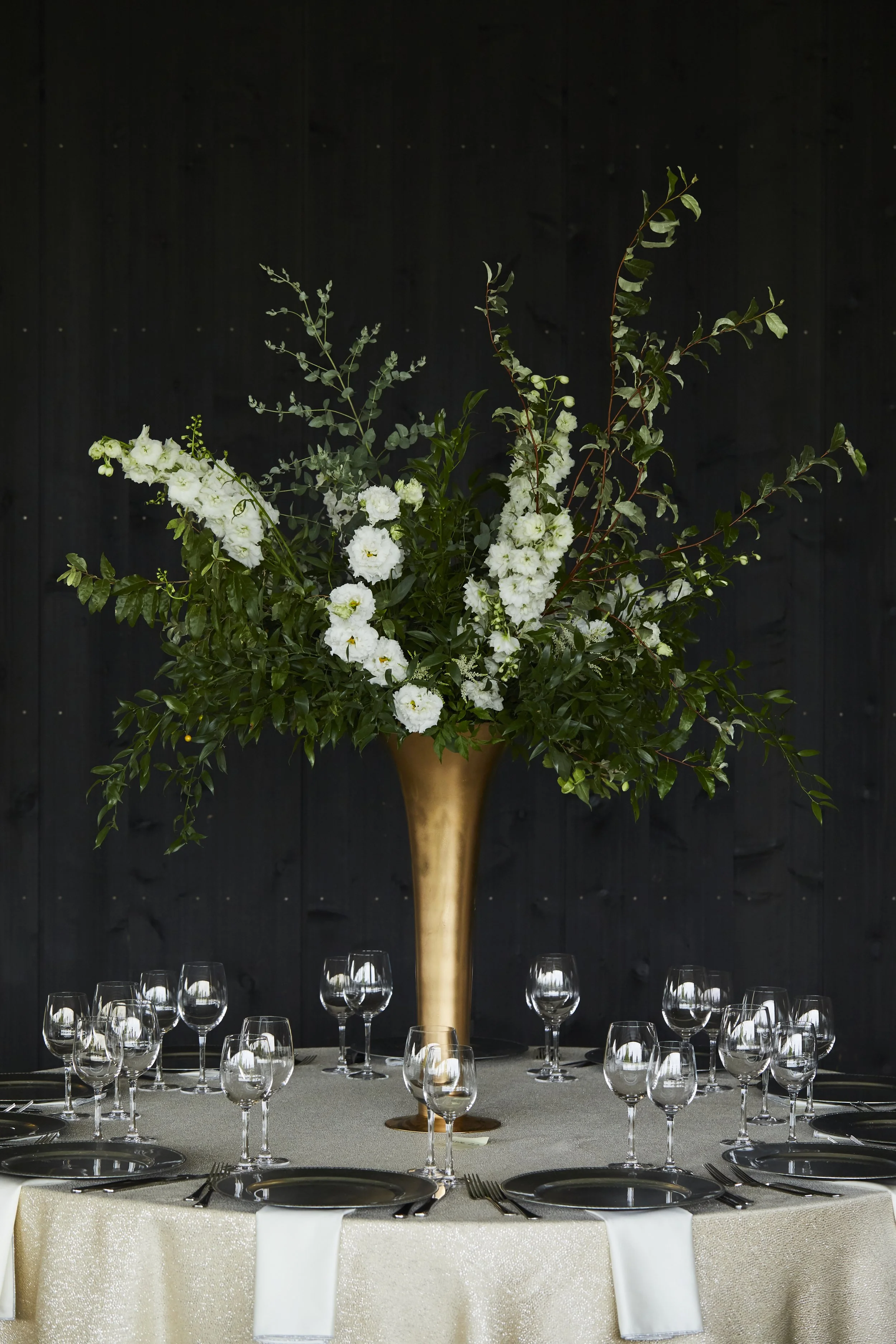 Elegant table setting with black plates, silver utensils, and empty wine glasses, centered with a tall floral arrangement of white flowers and greenery in a gold vase against a black backdrop.
