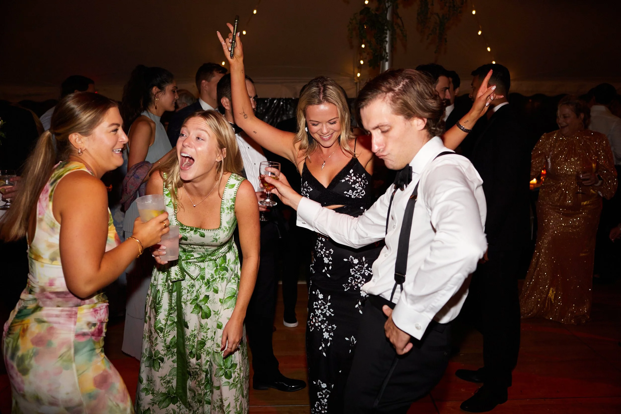People dancing and socializing at a lively party or celebration, with a woman in a black dress smiling and dancing, and others in elegant attire holding drinks.
