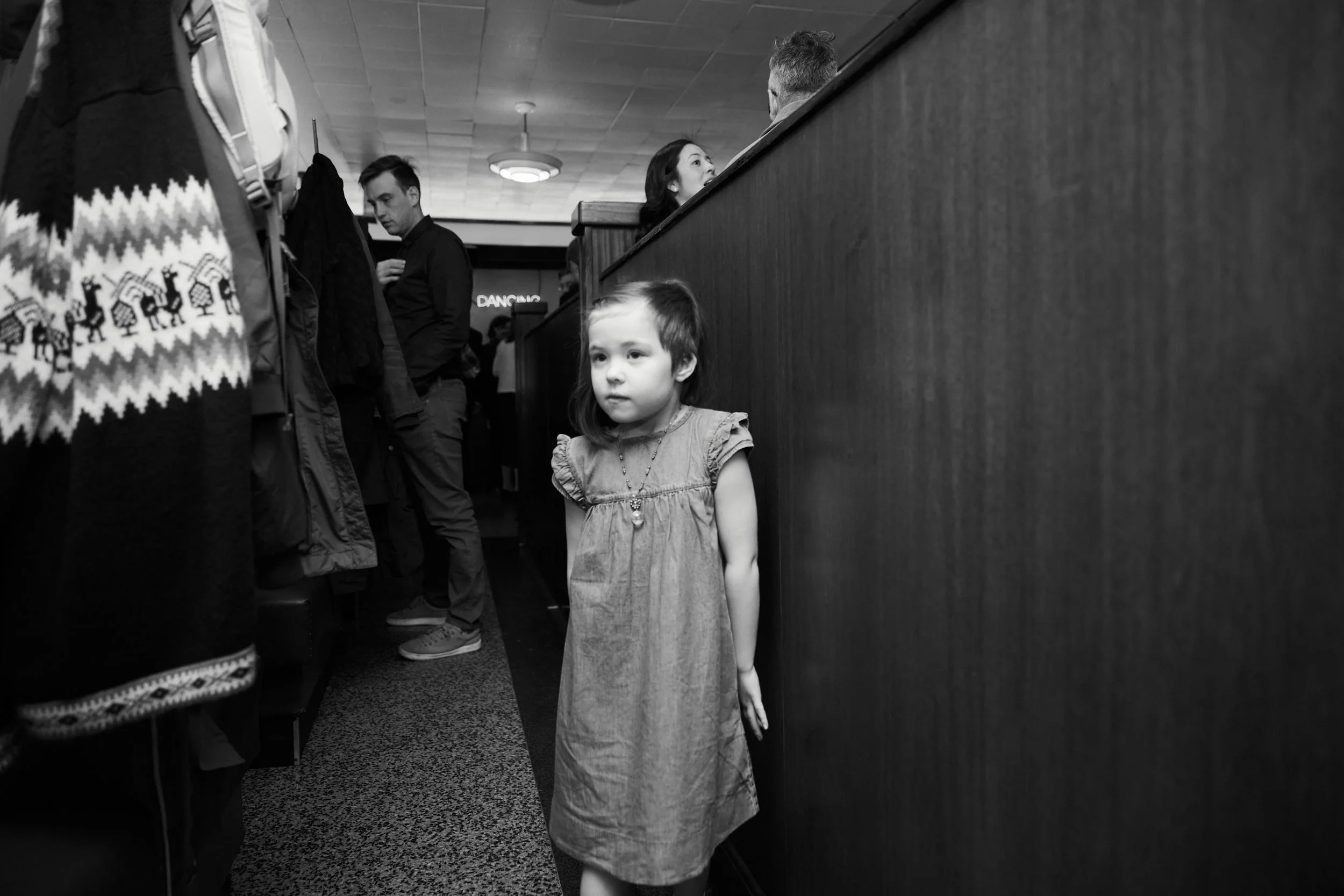 A young girl standing alone in a store aisle, looking ahead with a neutral expression, surrounded by other shoppers and clothing items.
