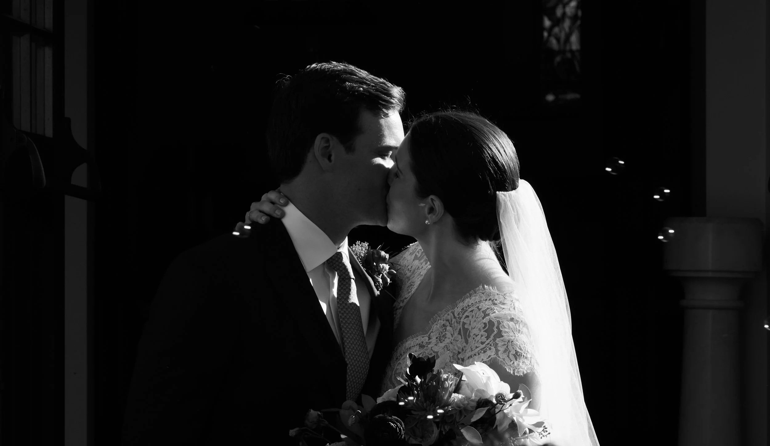 Black and white photo of a bride and groom kissing, with the bride holding a bouquet of flowers and wearing a lace wedding dress and veil.