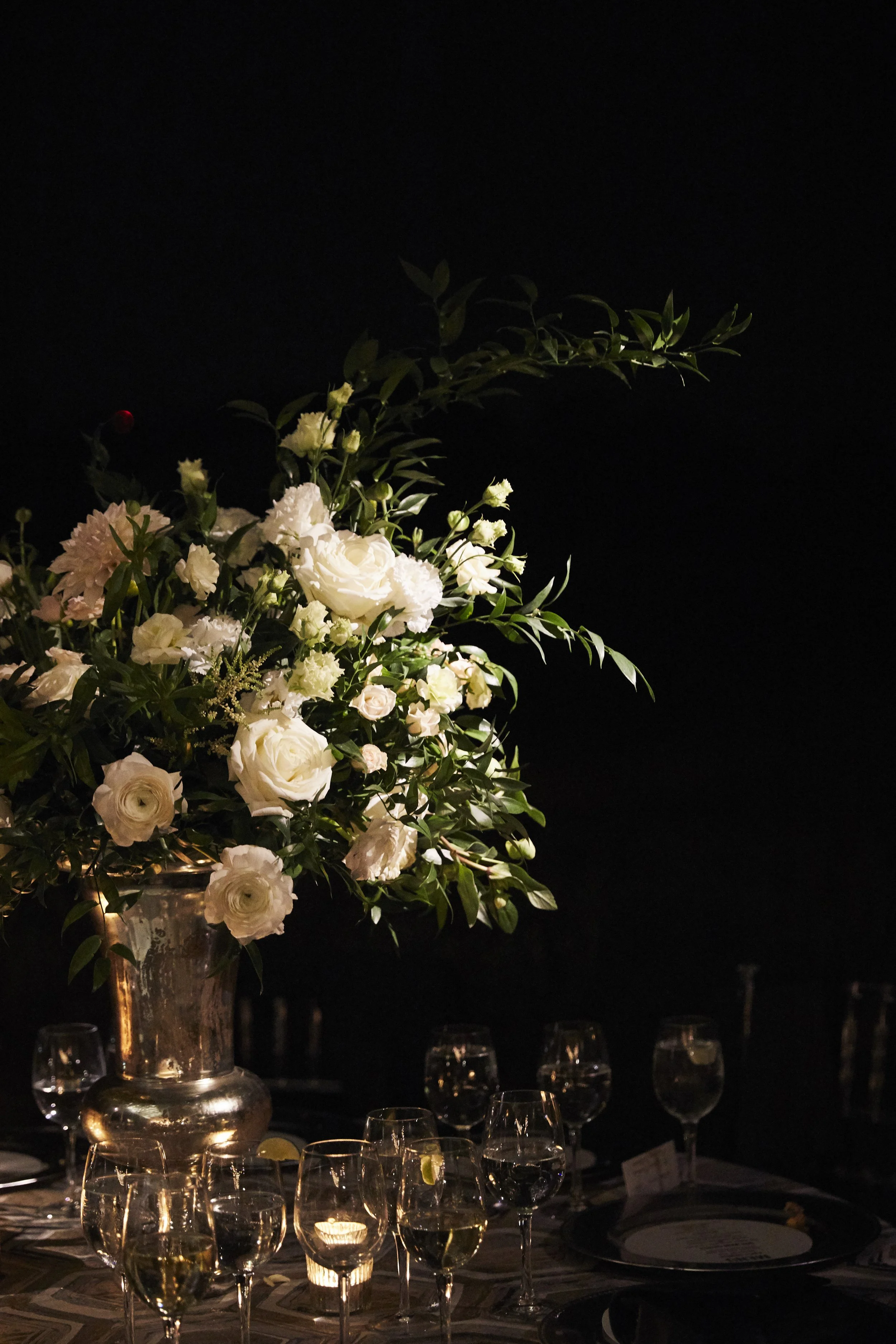 A large floral centerpiece with white and soft pink flowers and green leaves on a dark table with multiple wine glasses and candles.