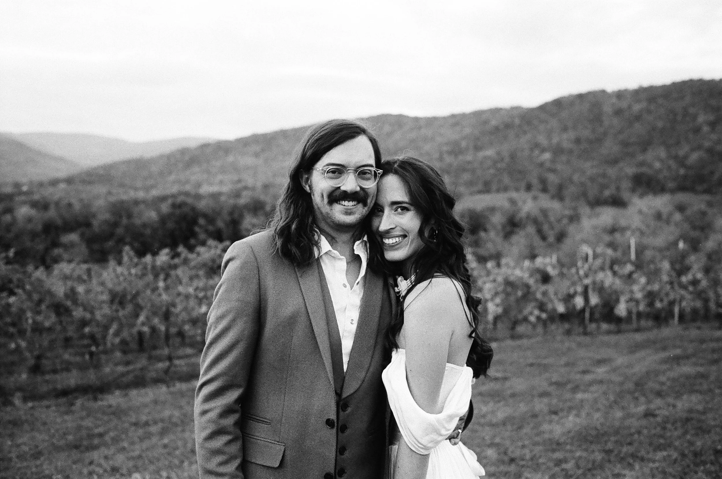 A man and woman smiling and standing close together outdoors with a mountain landscape in the background.