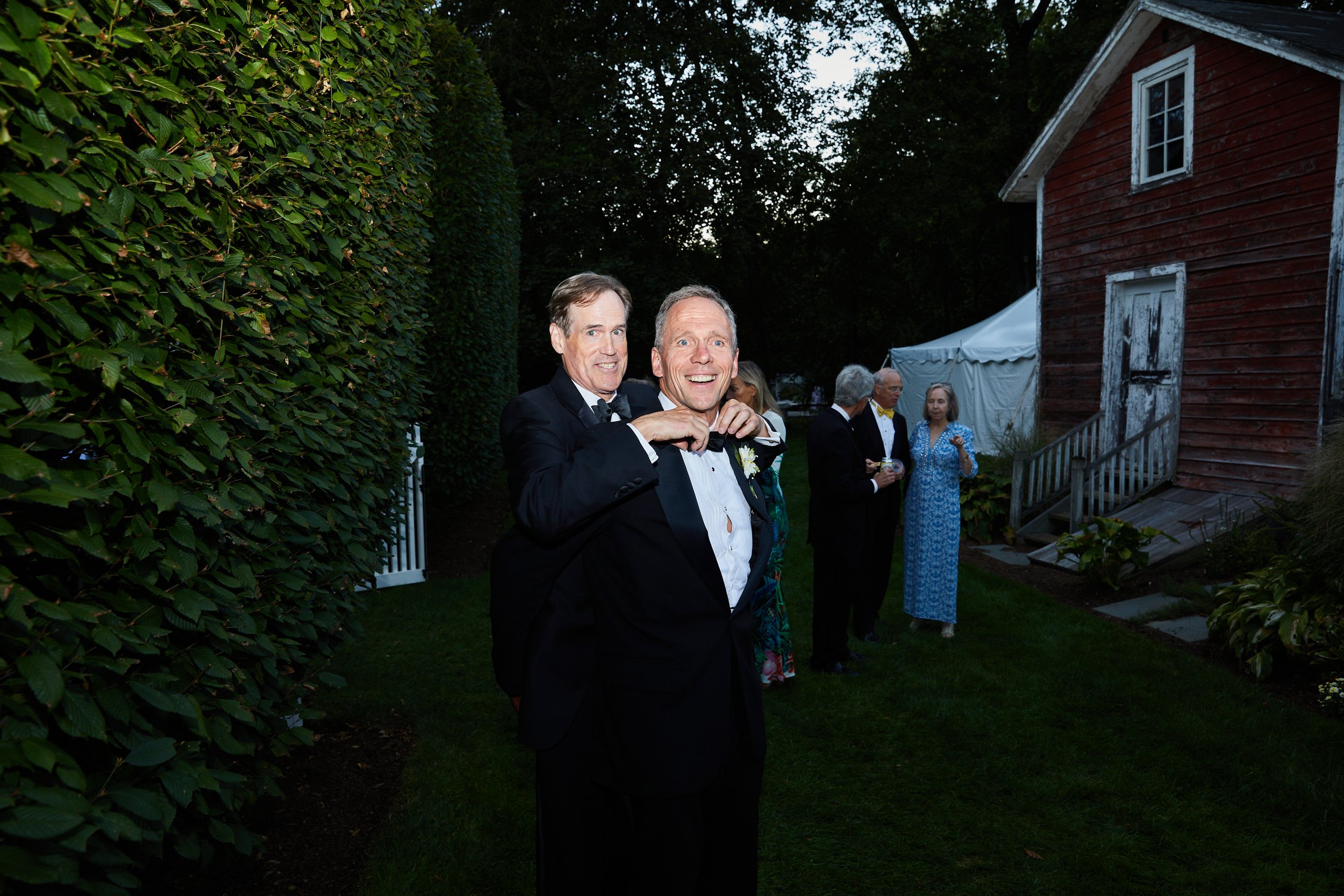 Two men wearing tuxedos at an outdoor evening event, with one man adjusting his bow tie while smiling at the camera; other guests in formal attire in the background near a red barn and white tent.