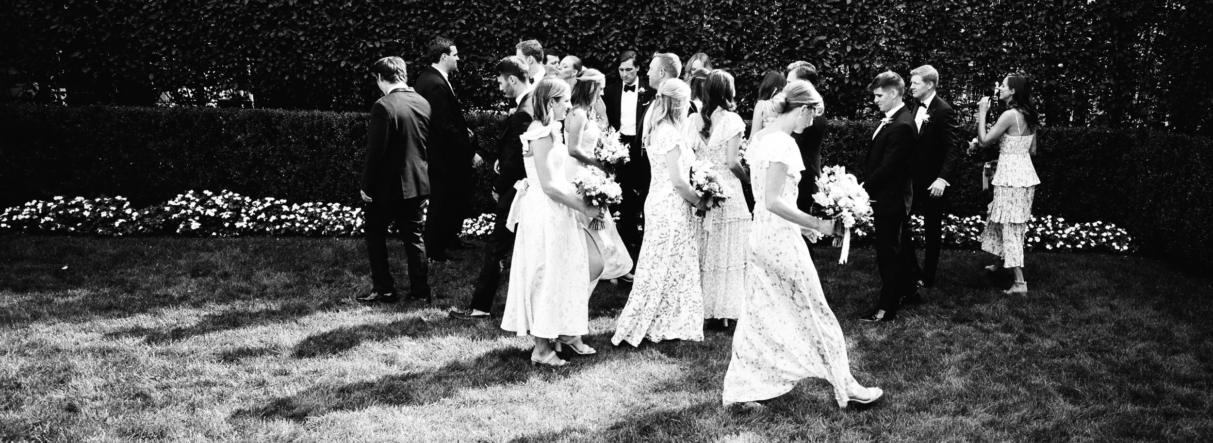Black and white photo of a wedding procession outdoors with brides and grooms walking on the grass, holding bouquets of flowers, with a hedge and flowers in the background.