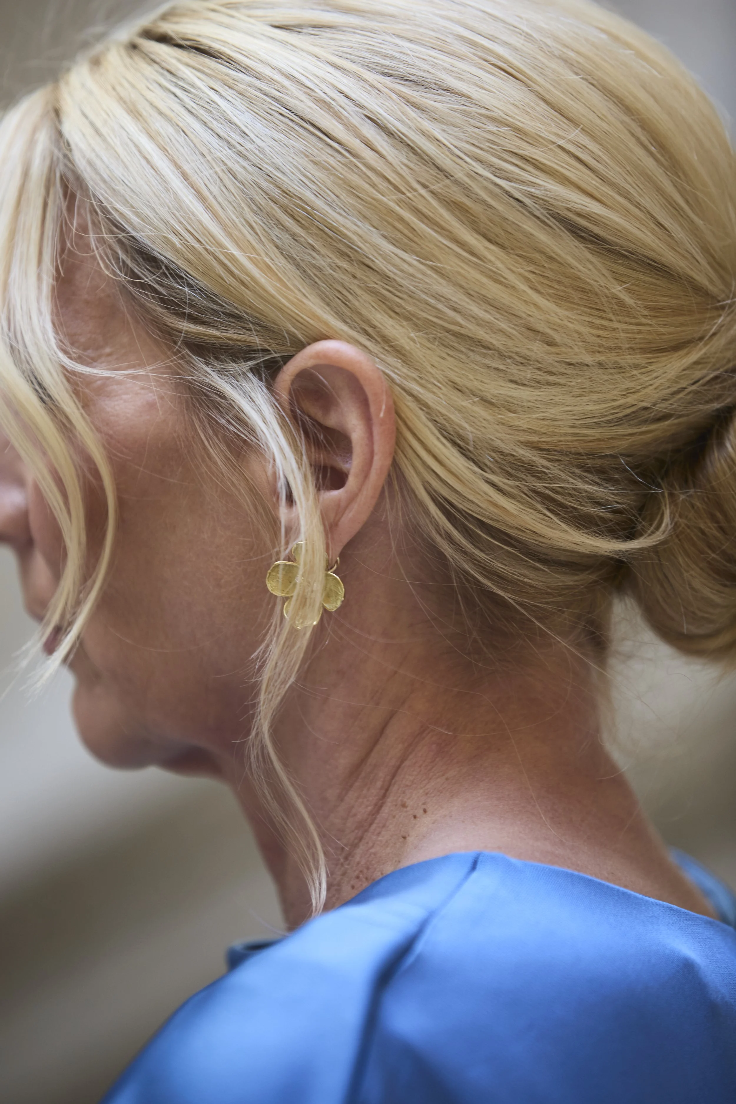Close-up of a woman's head, showing her blonde hair styled in a bun, with a gold earring and a blue garment.