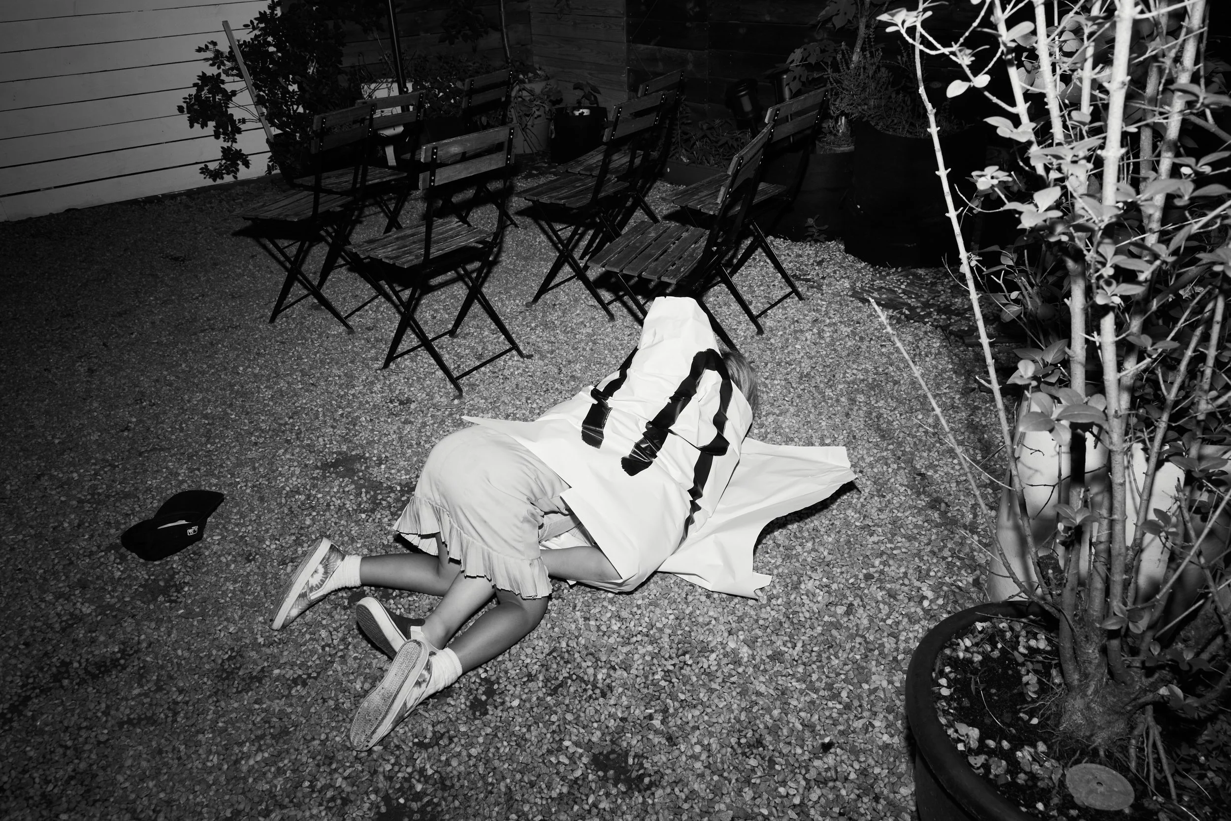 A child lying on the ground wrapped in a large white trash bag, with legs and shoes visible, in an outdoor patio area at night with tables, chairs, and plants.
