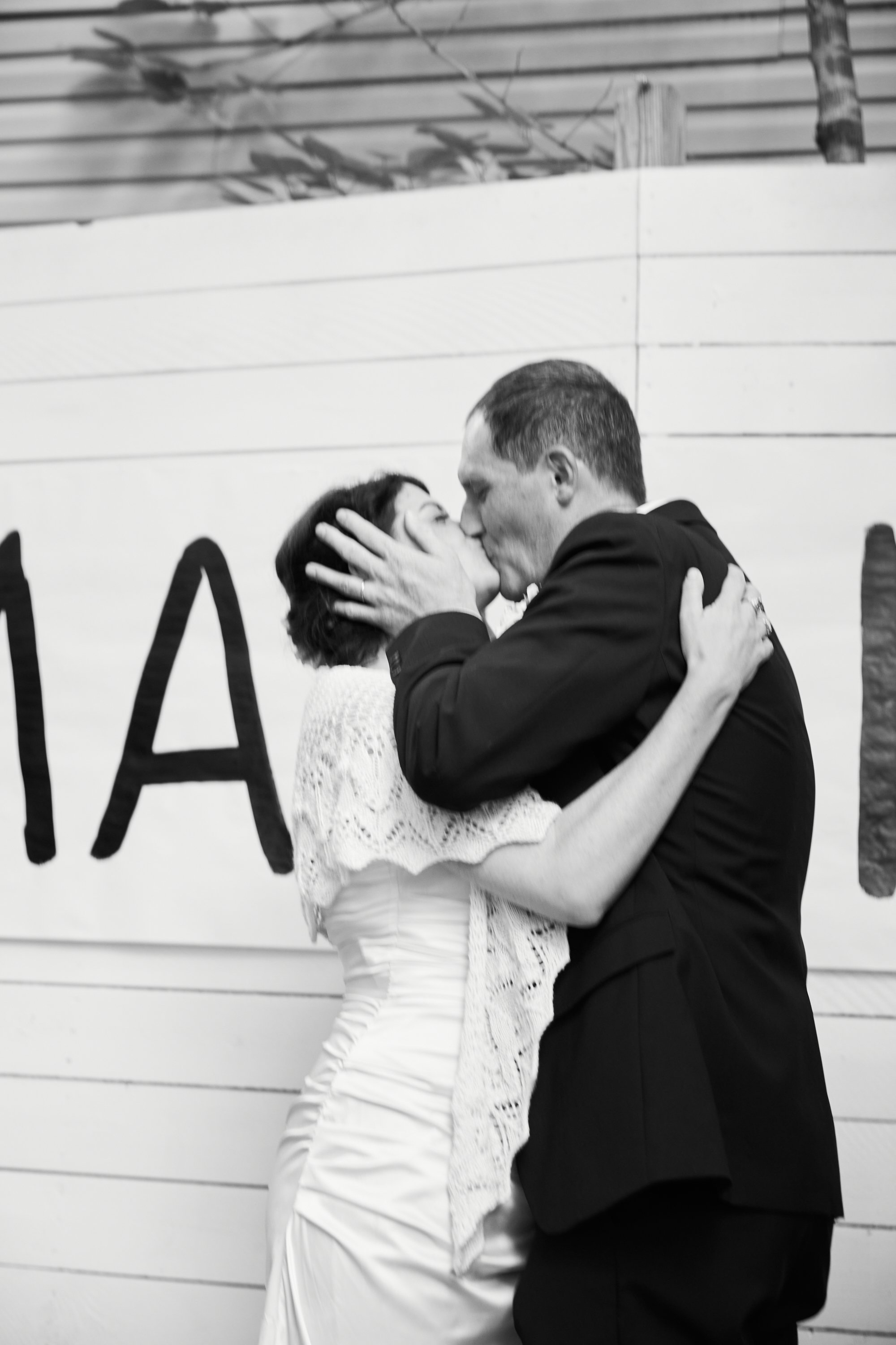 A couple in wedding attire sharing a kiss and embracing in front of a wall with a partial sign.