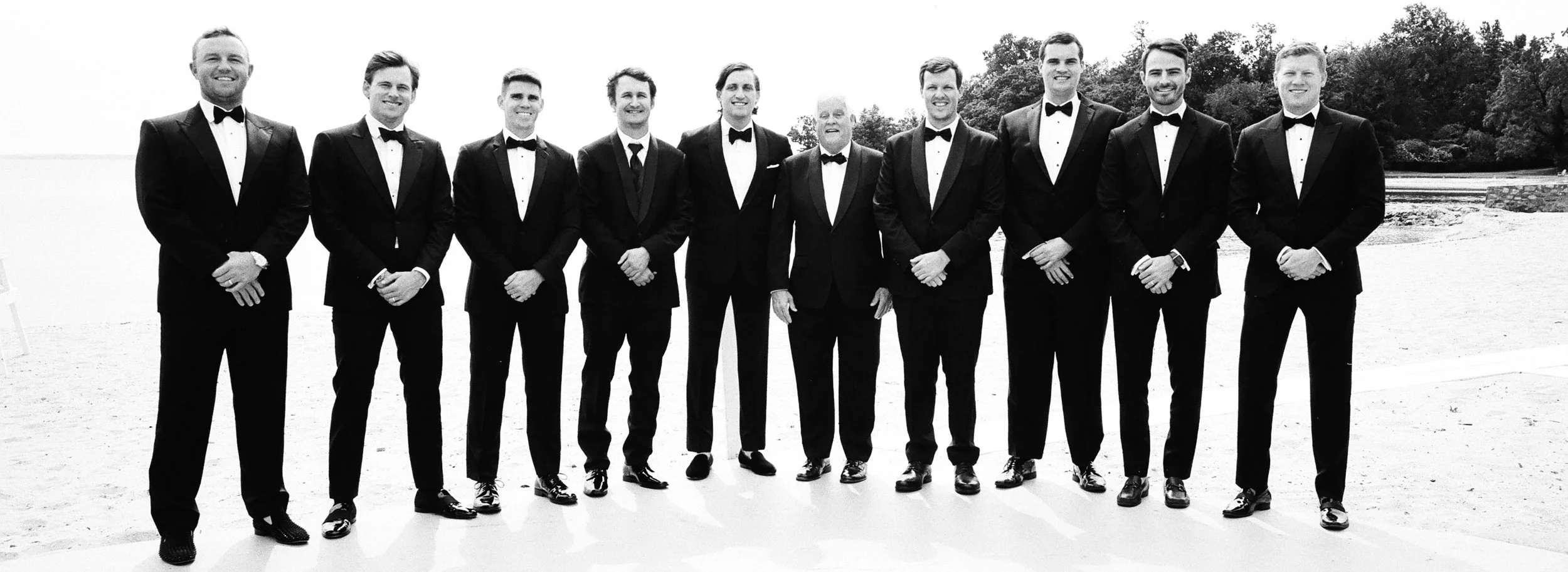 A group of eleven men dressed in tuxedos standing outdoors on a sandy beach with trees in the background.