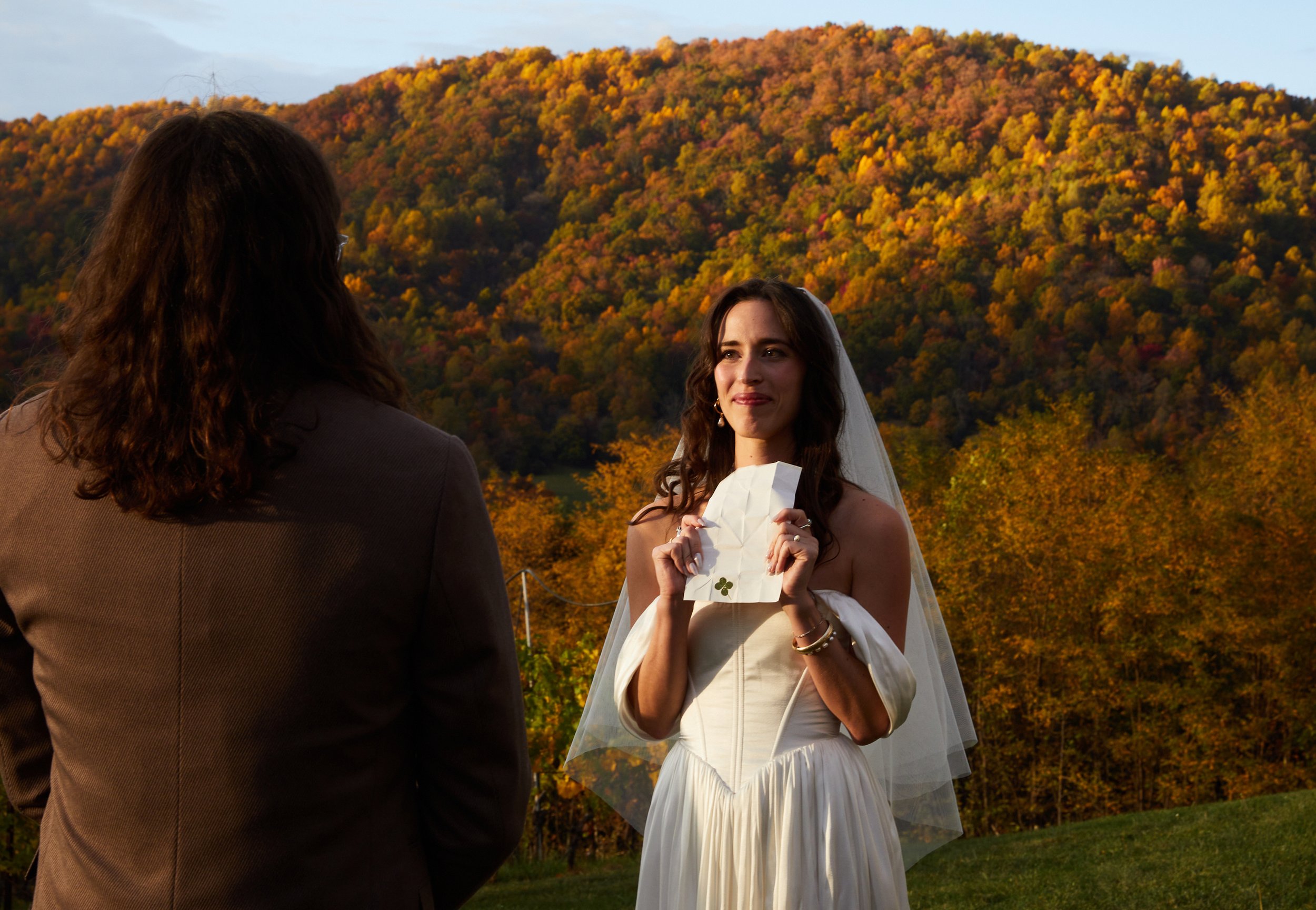 A woman in a wedding dress holding a letter with a four-leaf clover, standing outdoors with fall-colored trees and hills in the background, during sunset.