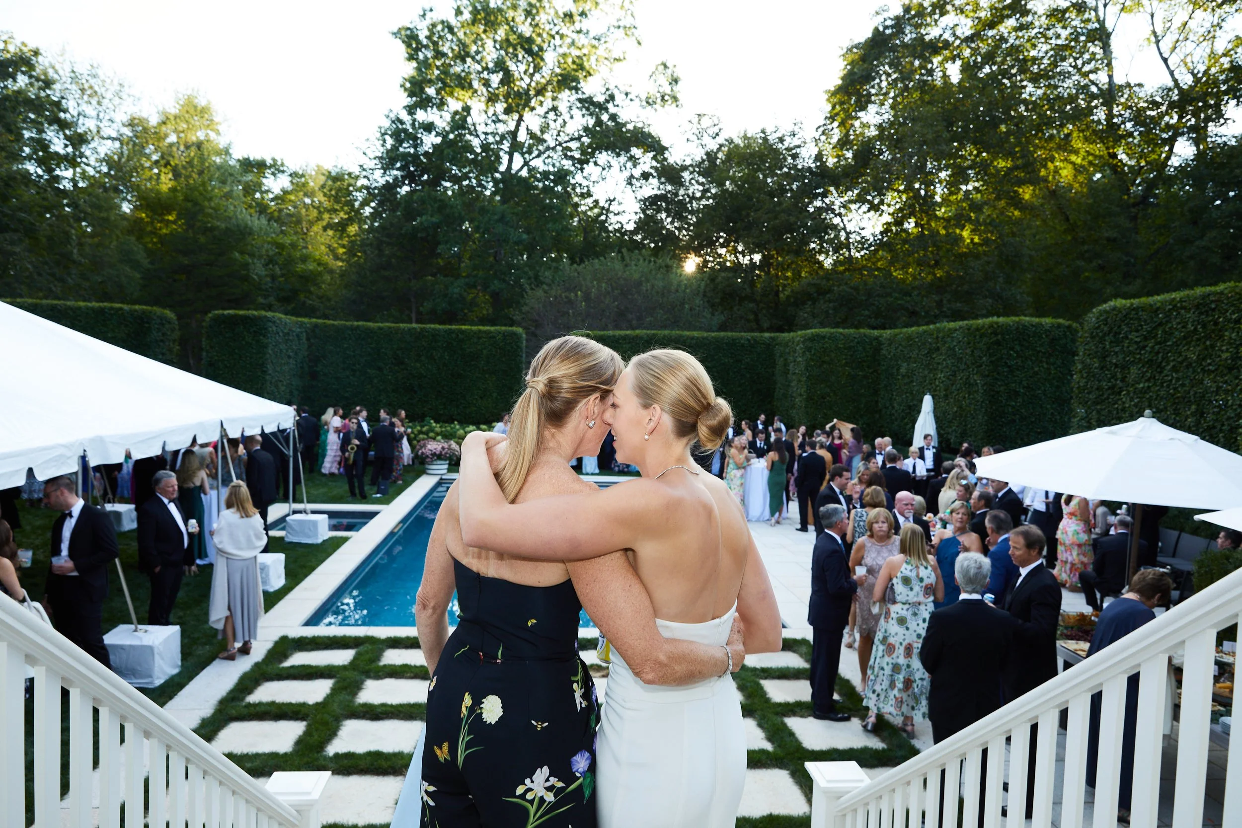 Two women, one in a black dress and one in a white dress, embrace and touch foreheads at an outdoor party with guests, a pool, and lush greenery in the background.
