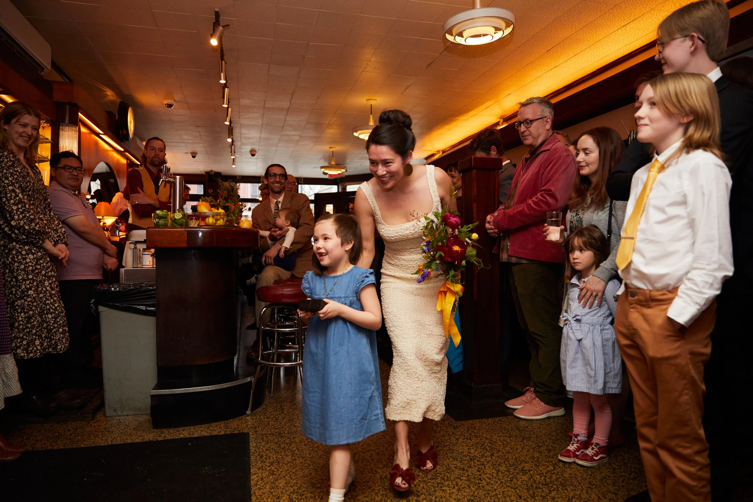 A woman in a white dress holding a colorful bouquet of flowers walks into a room filled with smiling people. The woman is accompanied by a young girl in a blue dress. Guests are standing along the wall, watching and smiling at the woman, with some ho