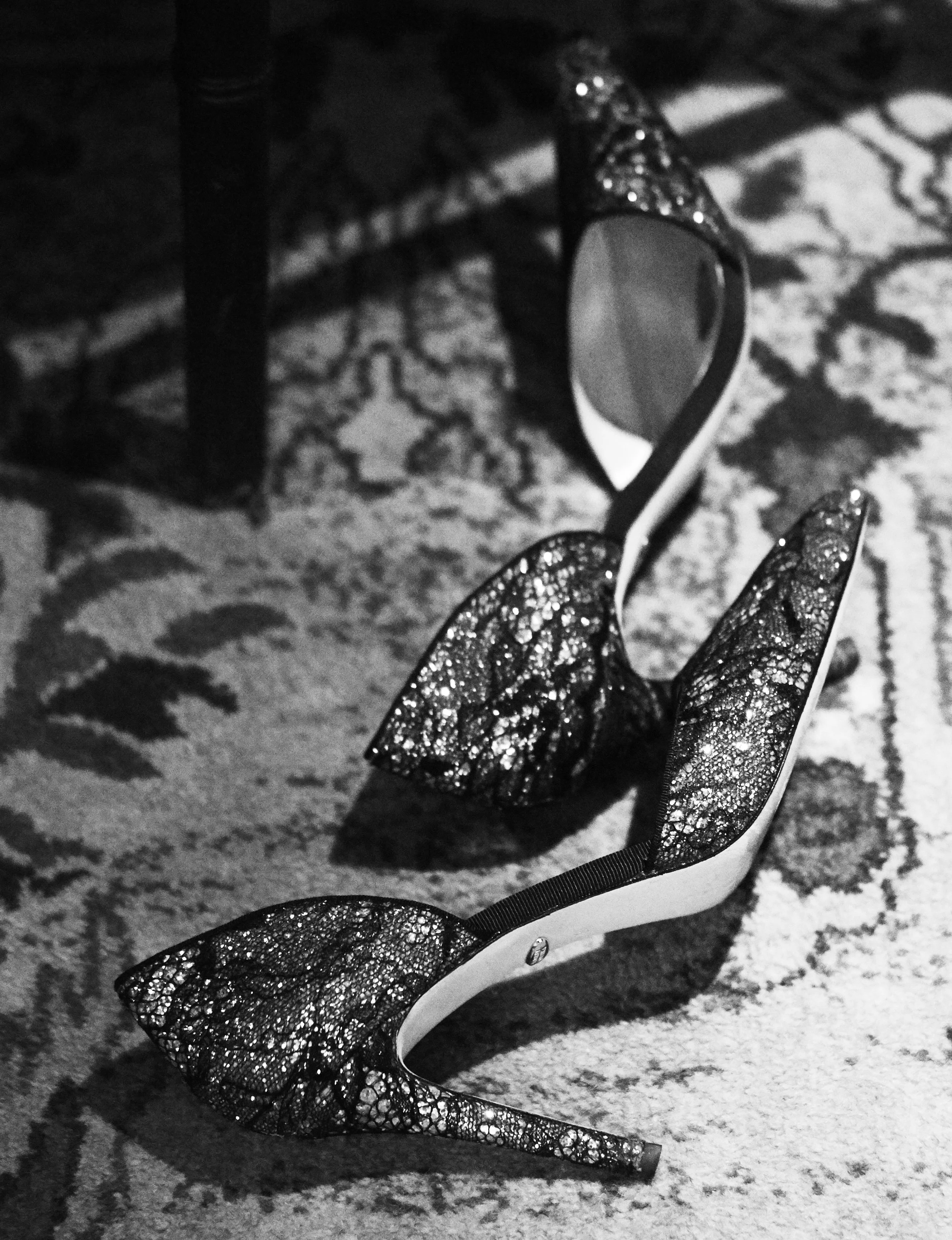 A high-heeled shoe placed on a patterned carpet, with decorative butterfly-shaped wings on the back of the shoe, featuring a glittery, lacy design.