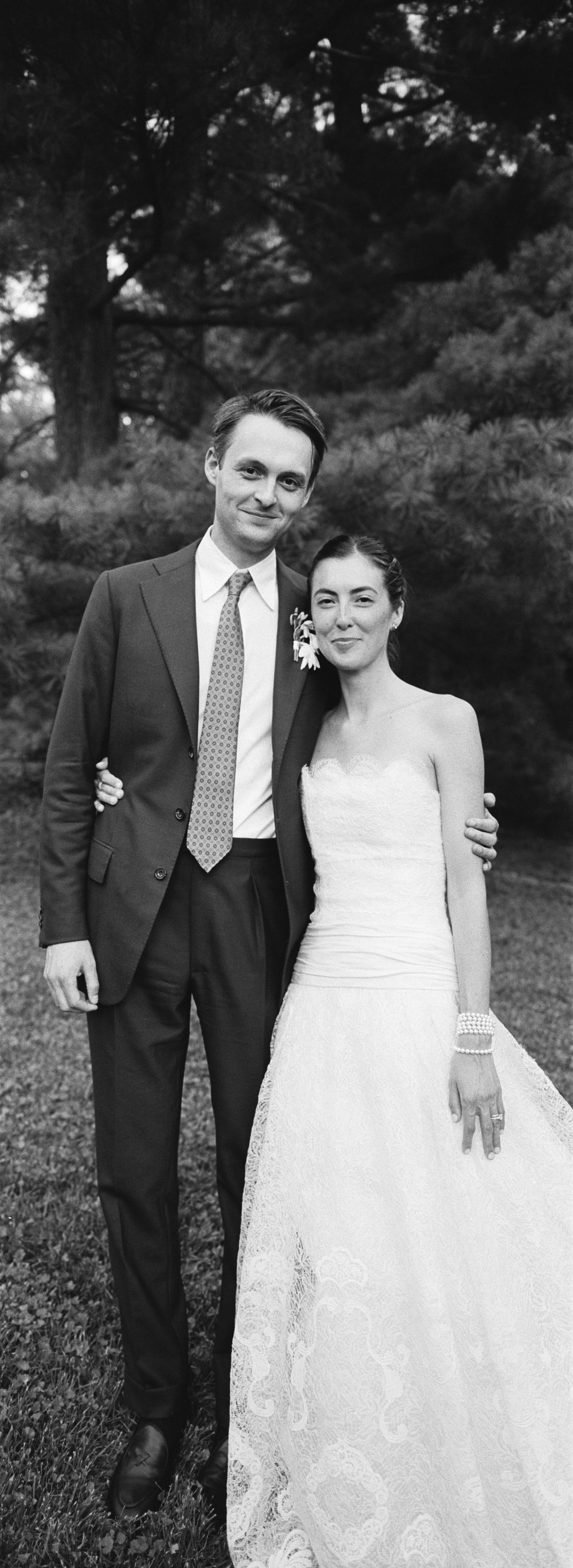 Black and white photo of a man in a suit and a woman in a wedding dress standing outdoors with trees in the background.
