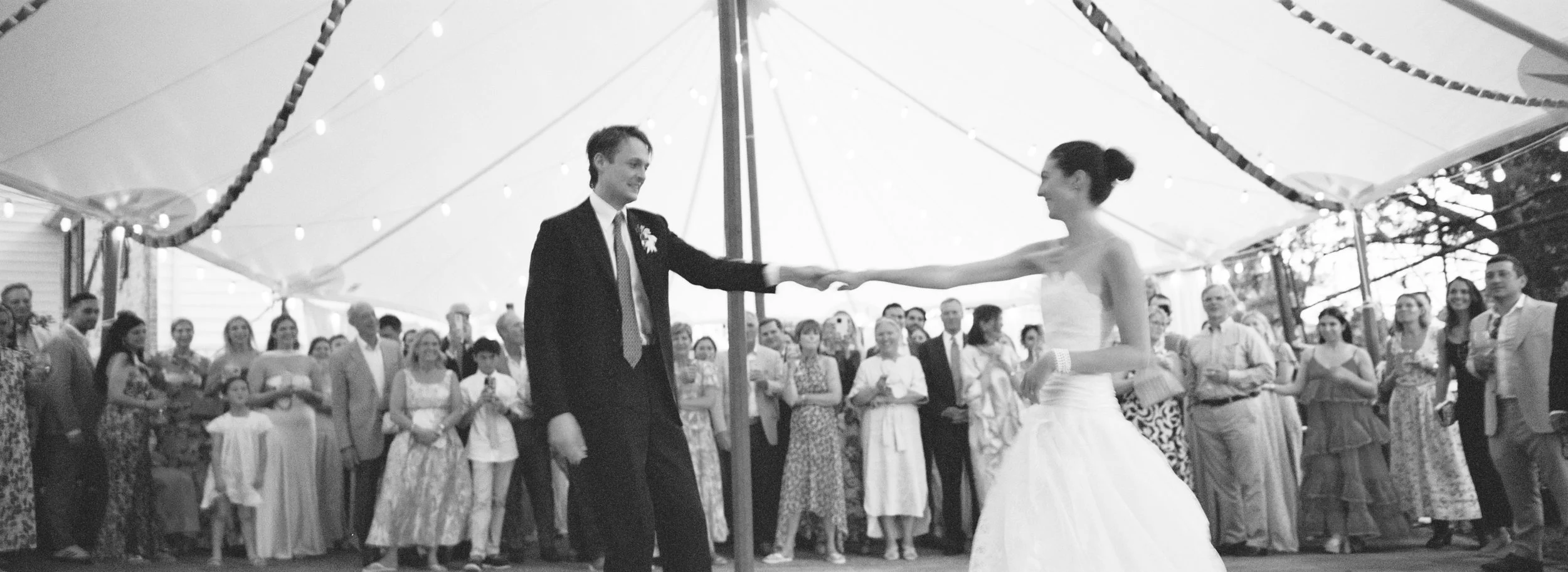 A black and white photo of a bride and groom dancing at their wedding reception under a canopy with string lights. The bride is wearing a strapless wedding gown, and the groom is in a dark suit with a tie. Guests are watching and smiling in the backg