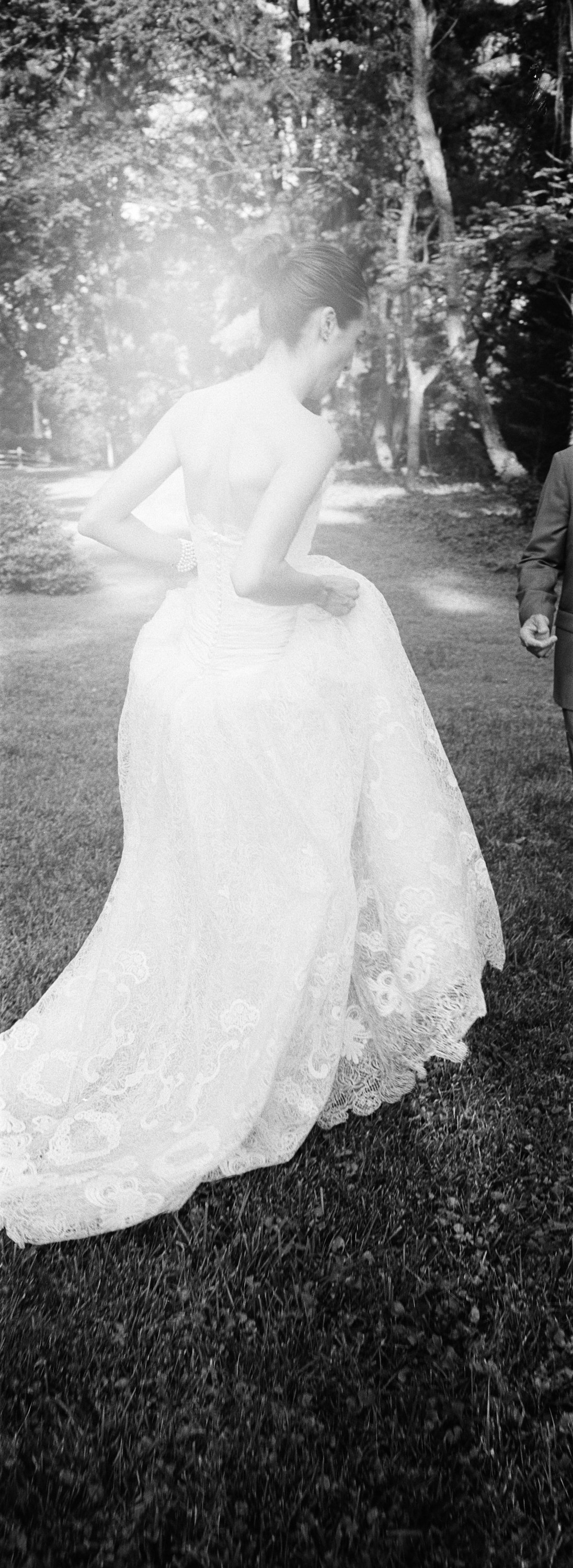Black and white photo of a woman in a wedding dress holding up the skirt, outdoors with trees in the background.